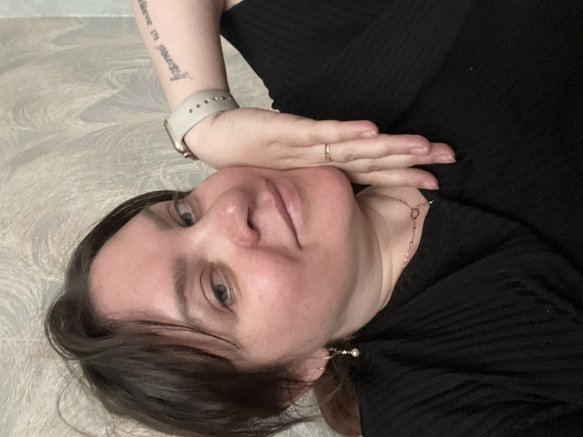 A woman with fair skin and dark hair is lying down, facing the camera. Her left hand is raised to her face, with her fingers resting near her chest. She is wearing a black ribbed top and has a light-colored watch on her left wrist. A gold ring is visible on her left hand, and a gold earring hangs from her left ear. A delicate gold necklace is also visible around her neck. A tattoo is seen on her left forearm. The background appears to be a light-colored surface with a textured pattern. The image was taken in Noviy, Russia.
