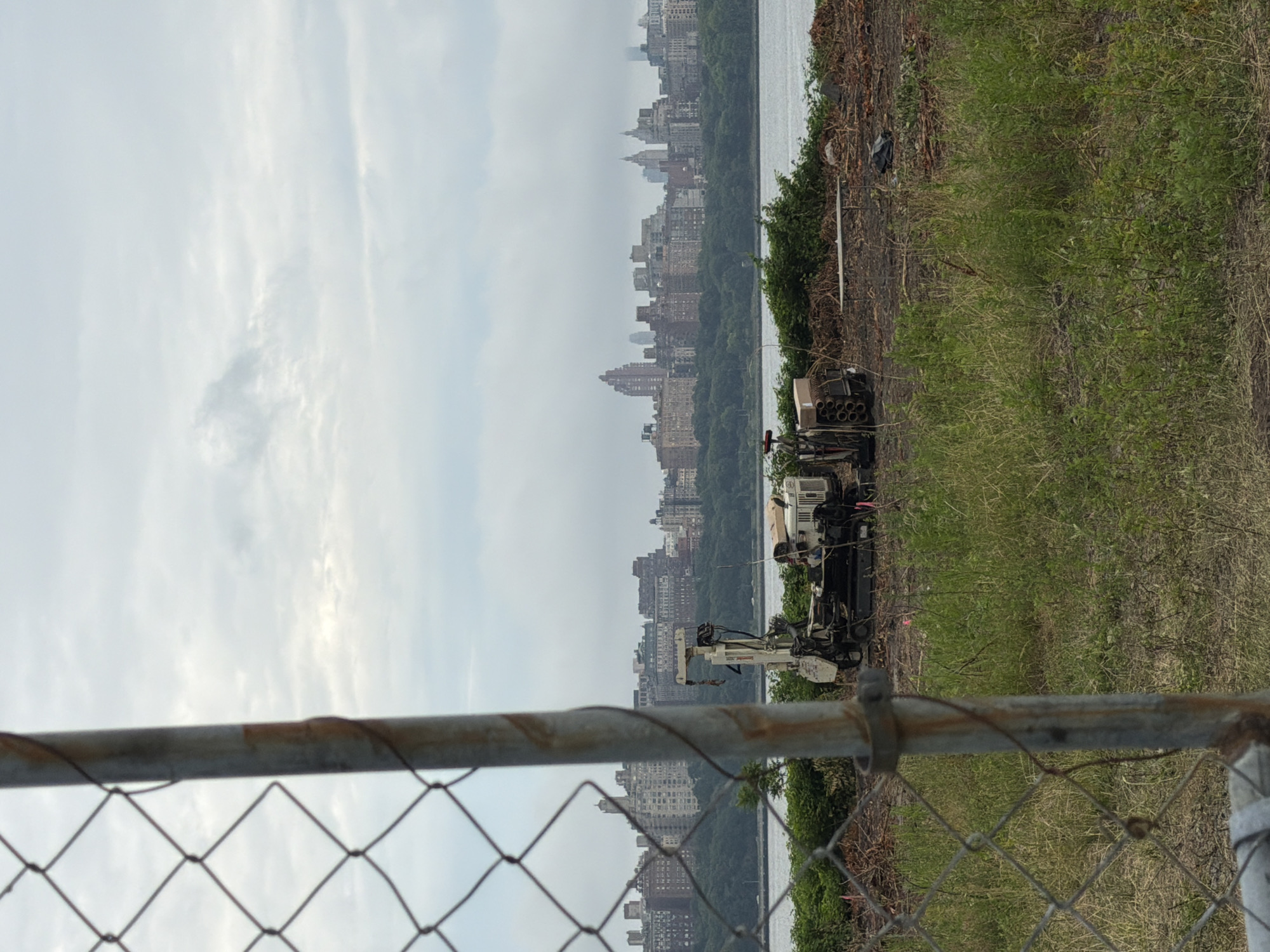 The image depicts an outdoor scene with a city skyline in the background, partially obscured by trees and a body of water. In the foreground, a chain-link fence runs horizontally across the bottom of the frame. To the right of the water and behind the fence, there is a patch of overgrown green grass and some dry brush.
On the right side of the image, a piece of heavy machinery, possibly a drilling rig or a construction vehicle, is parked on a patch of dirt and debris. The machine appears to be white and black with various components and a boom. Next to it, there is a cardboard box and what looks like a stack of cylindrical objects.
The skyline in the background features numerous buildings, suggesting an urban environment. The sky is overcast, with soft, diffused light, indicating it is likely daytime, perhaps late morning or early afternoon, under cloudy conditions. The overall impression is of an industrial or construction site located near a city and a body of water. There are no people visible in the image. No visible text is present.