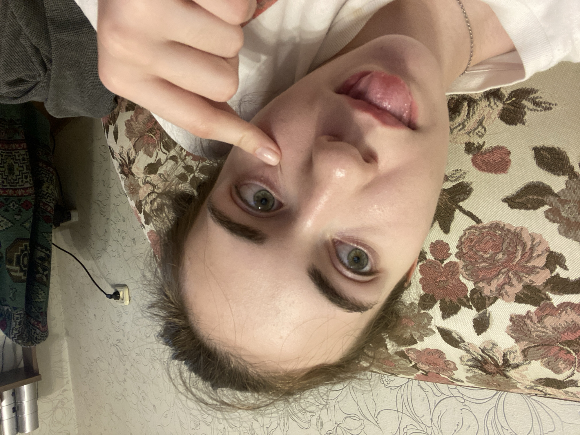 A young woman with light skin and green eyes is lying down, partially visible on a floral patterned sofa. She is looking at the camera and sticking her tongue out. Her right index finger is touching her cheek near her eye. The background consists of patterned wallpaper and a wall outlet with a cord plugged into it. A shelf with cylindrical objects and a draped fabric can be seen on the right side of the image. The scene appears to be indoors, likely in a residential setting. There is no information in the image to determine the location within the city of Zavodoukovsk, Russia.