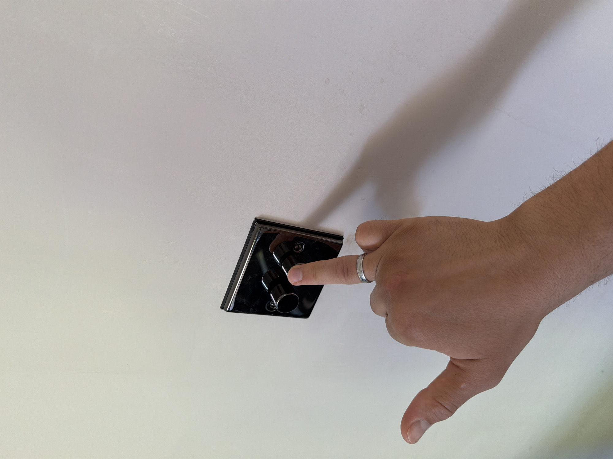 In a brightly lit room, a person's left hand is positioned near a black, square-shaped light switch plate mounted on a textured white wall. The switch plate has two black, cylindrical knobs. The index finger of the hand is pressing down on the right knob, and a silver band is visible on the ring finger. The thumb is extended, and the hand is casting a shadow on the wall. The background is a plain white wall, with some subtle textures and faint marks. The location in Leeds, United Kingdom is not specified by the image.