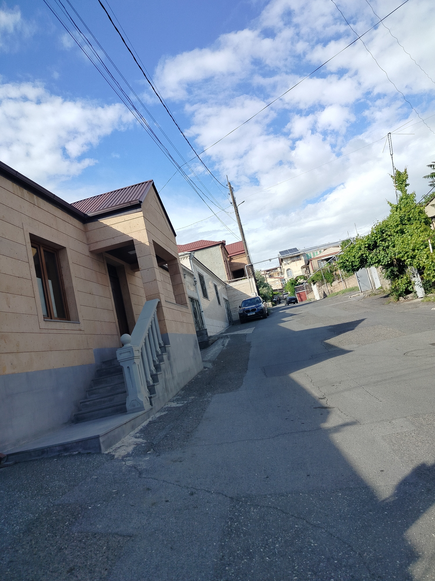 The image shows a residential street in Yerevan, Armenia, captured on a sunny day with scattered clouds. The road slopes slightly upward and is paved with asphalt. On the left side of the street is a modern, single-story house with light-colored stone cladding, a brown roof, and a short staircase leading to the entrance. The house's exterior includes a window and a partially shaded doorway. A section of a neighboring white building is visible behind the house.
Further up the street, on the left, is a dark-colored car parked near another building. On the right side of the street, there is dense green vegetation and a fenced area. Utility poles and wires stretch across the sky above the street. Other houses and buildings are visible further up the street, suggesting a residential neighborhood setting. The shadow of the photographer is cast on the road in the foreground.