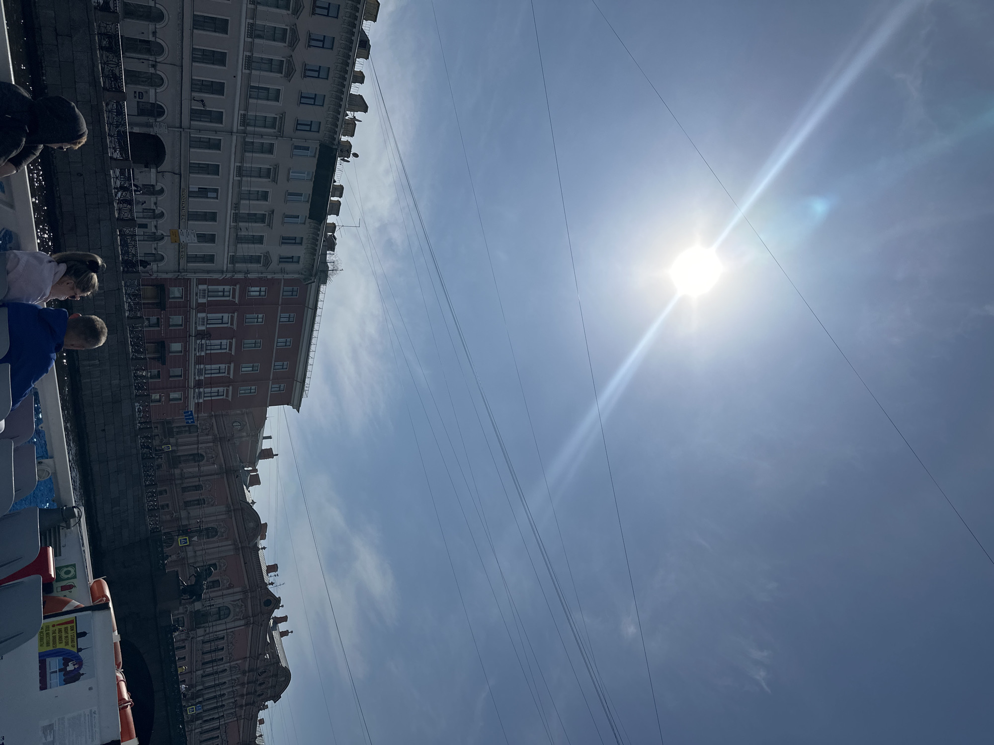 The image captures a scene in Saint Petersburg, Russia, as viewed from a boat on a canal. The perspective is angled upwards towards the sky, where the sun shines brightly, causing a lens flare. Buildings line the canal edge, exhibiting a mix of architectural styles and varying heights. A group of people are partially visible on the left side of the image, presumably passengers on the boat. Electrical wires crisscross the sky, adding to the urban landscape. A sign with the words "DON'T STAND UP RIGHT BEFORE AND UNDER THE BRIDGEST" is visible, indicating the canal is navigable by boats with low headroom. The text "1292295", "ПРОДУКТЫ 24" and "ВСТАВАТЫ" is also seen on the left side of the image.