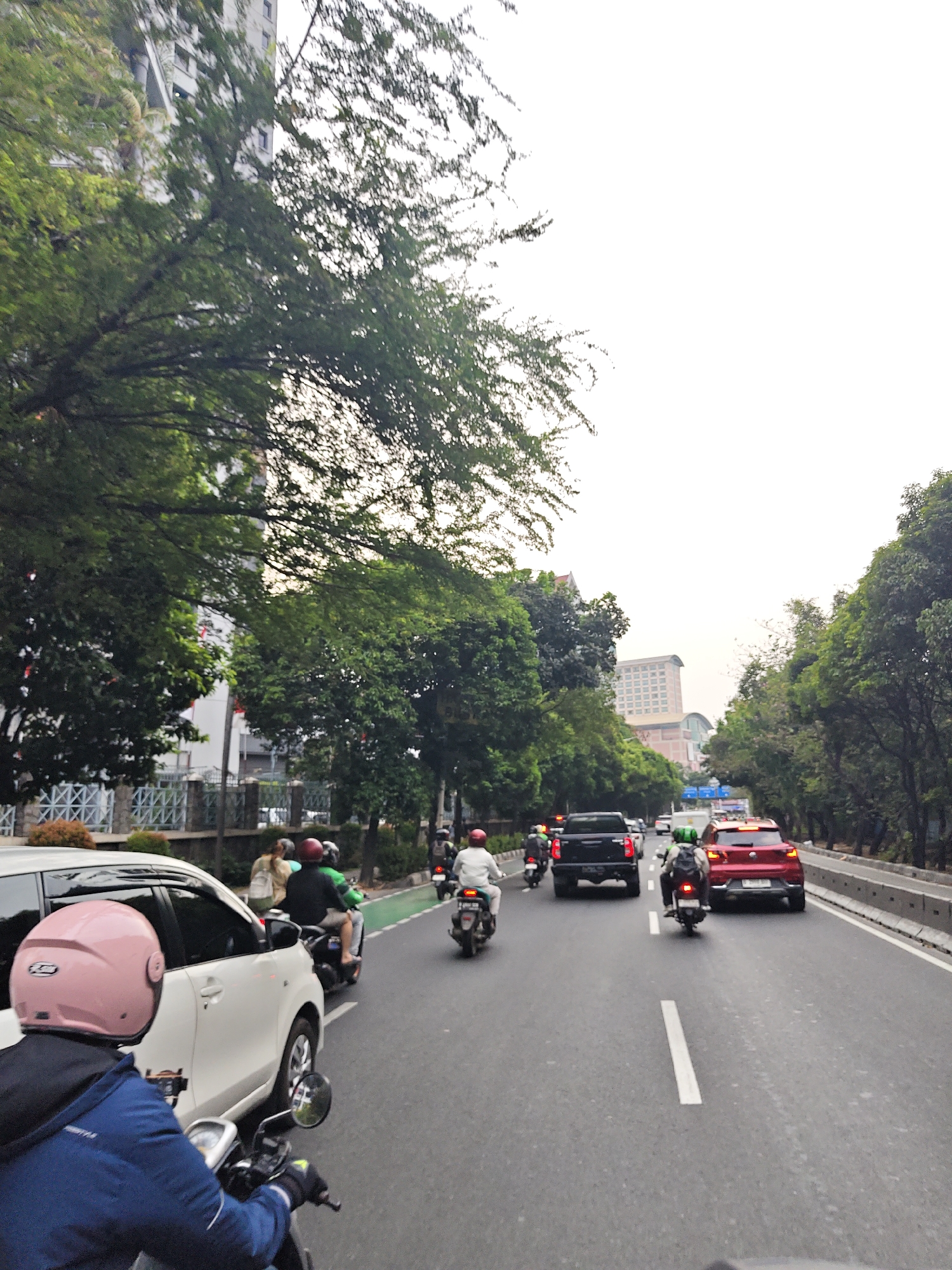 This image captures a street scene in West Jakarta, Indonesia, characterized by moderate traffic under an overcast sky. The perspective is from the back of a motorcycle, looking forward down a multi-lane road bordered by lush green trees and buildings.
In the foreground, a person wearing a blue jacket and a pink helmet is riding a motorcycle. Beside them, a white car is also in motion. Further down the road, several other motorcycles and vehicles are visible. One motorcycle has a rider in a green helmet with a backpack, and another has a rider in a red helmet. A black pickup truck is in the middle lane, followed by a red SUV.
The environment includes a concrete barrier separating the road from the sidewalk, where more trees and some pedestrian activity can be observed. In the background, tall buildings with multiple stories are present, suggesting an urban setting. The overall atmosphere appears to be one of daily commute or travel within the city.