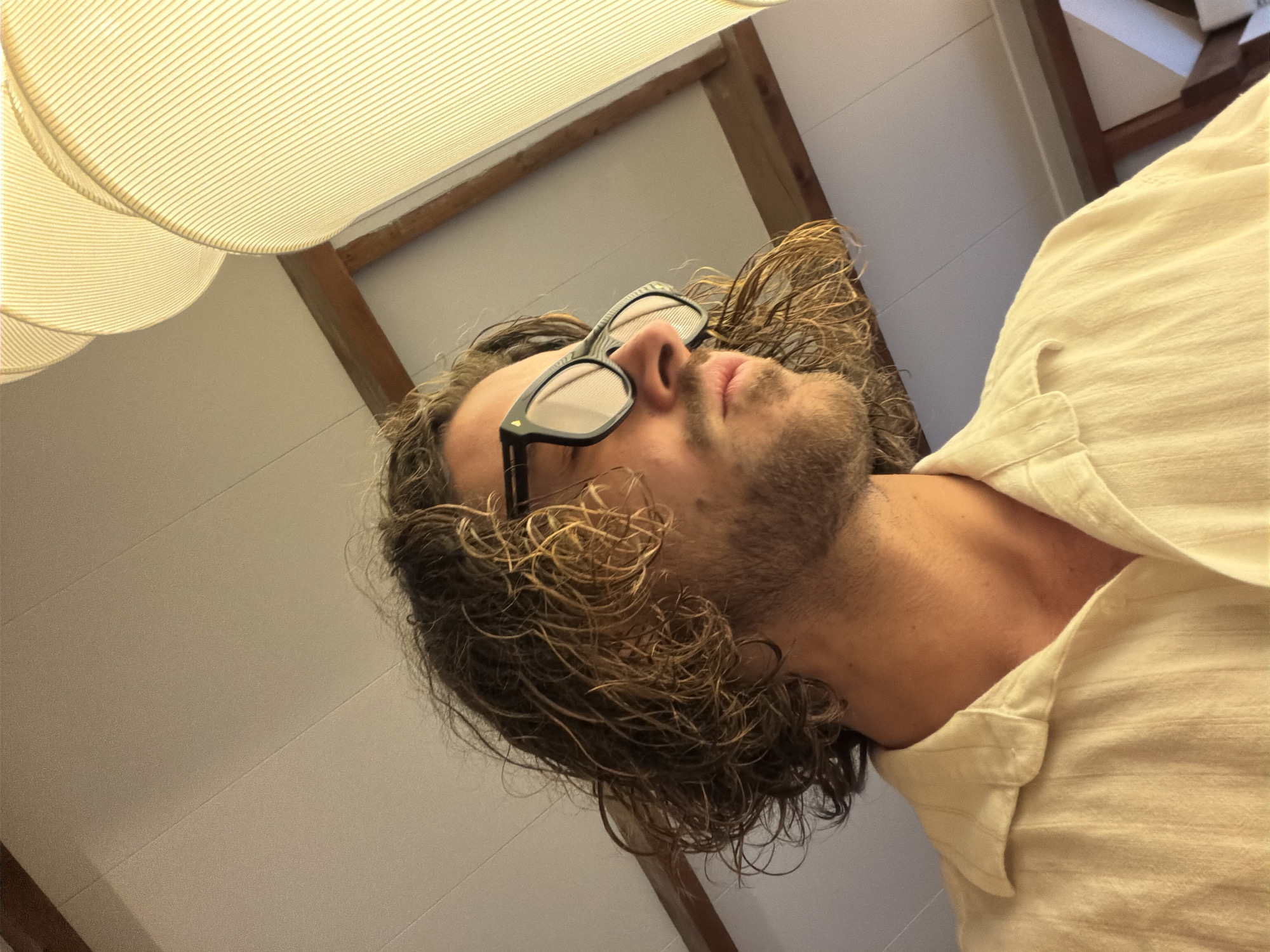 This media file presents a low-angle portrait of a man, likely in an indoor setting in Paris, France. The subject, seen from the chest up, is looking upwards with a serene or contemplative expression, obscured by dark-framed sunglasses resting on his nose. He has medium-length, curly brown hair and a light beard or stubble. He is dressed in a light-colored, possibly cream or off-white, collared shirt with visible texture.
The background primarily features a light-colored, horizontally paneled ceiling, along with exposed wooden beams that add to the architectural character of the space. Dominating the upper left portion of the frame are two distinct light fixtures with ribbed, semi-transparent shades, which emit a warm, diffused glow, suggesting ambient lighting. The overall atmosphere conveyed by the warm light and the man's relaxed posture is one of calm or repose. There is no visible text in the image.