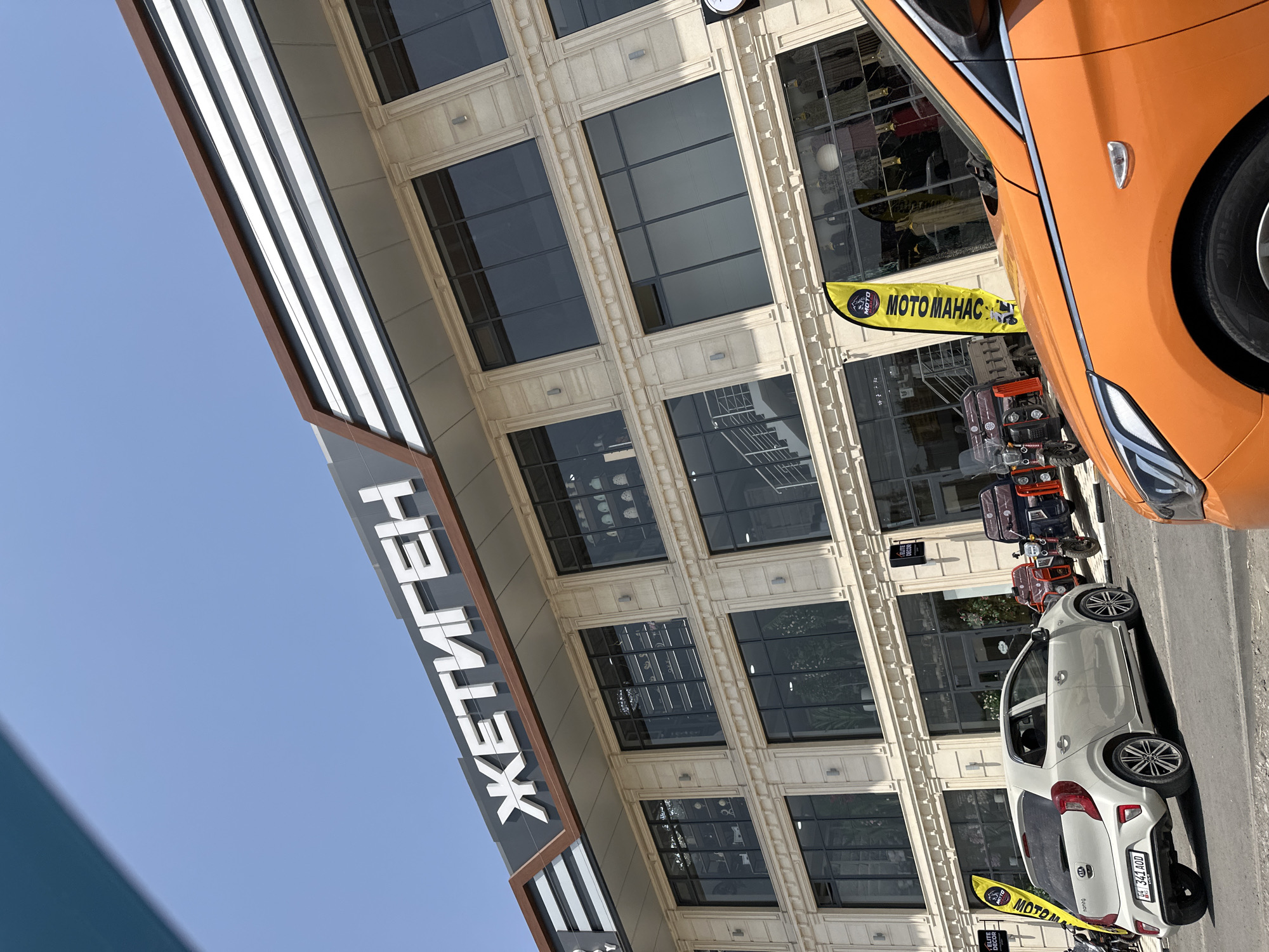 The image shows a modern, multi-story building with large windows and ornate architectural details, likely a commercial establishment. The building has a prominent sign with Cyrillic lettering that reads "ЖЕТИГЕН" in large, white, raised letters against a dark background. The building is lit by natural sunlight under a clear blue sky. In the foreground, several vehicles are parked, including a white Kia Picanto with a license plate visible, and the front of an orange car. Behind the white car, there are three-wheeled vehicles that appear to be electric scooters or small utility vehicles. Yellow flags with graphics and text are placed near the entrance of the building. Inside the windows, various items are displayed, suggesting retail shops. The overall scene depicts a commercial area on a sunny day in the city of Manas, Kyrgyzstan.