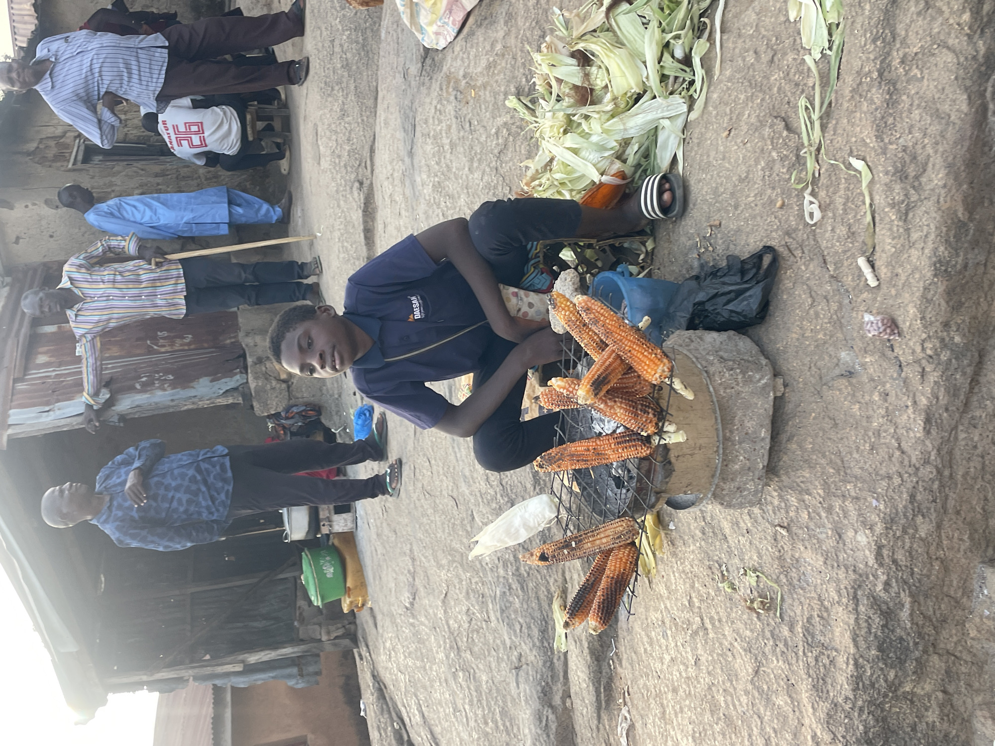 A young man in a dark blue short-sleeved shirt with the word "DAESAM" visible on the chest sits on a rock surface and tends to a grill with corn cobs on it. Some cobs are cooked and brown, others are white and uncooked. Corn husks are scattered nearby. He wears dark pants and striped sandals. Several people, including a man in a light blue shirt, are standing or walking in the background near structures with corrugated metal roofing. The setting appears to be in a public area. The location is Jos, Nigeria.