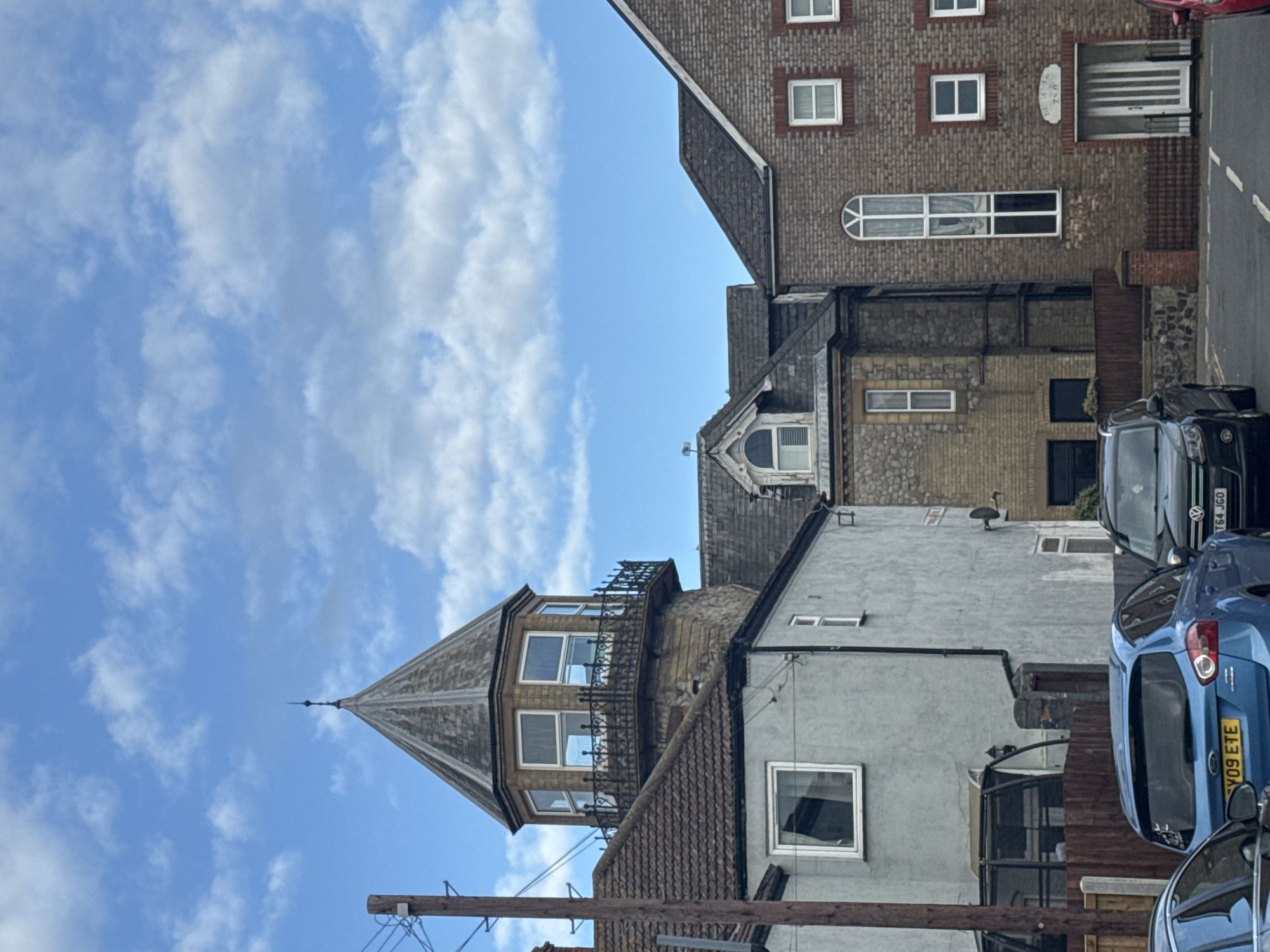 The image depicts a street scene in Gorleston-on-Sea, United Kingdom, angled upward to capture the architecture and sky. Several buildings of varied styles are visible, including brick and stone constructions. The tallest building features a pointed turret with decorative ironwork and windows. A blue car is parked on the right side of the frame, with a partially visible license plate reading "Y09 ETE". Other cars are also visible in the background. Power lines and utility poles are in the lower part of the image. The sky is blue with scattered white clouds.