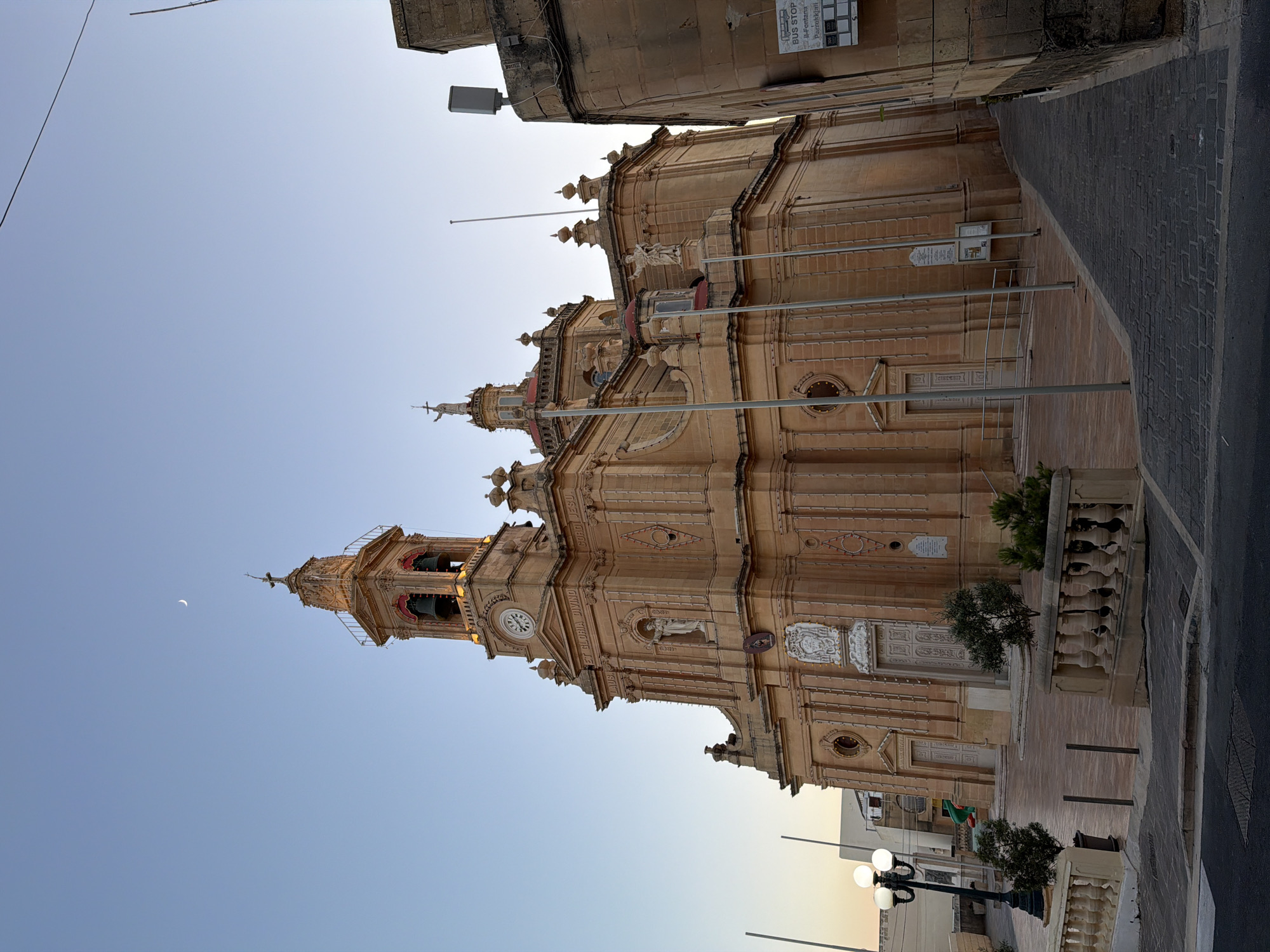 The image depicts the exterior of the Marija Bambina Church in Fontana, Malta, during what appears to be sunset or early morning due to the soft lighting and the crescent moon visible in the clear sky. The church, constructed from a light-colored stone, features a prominent bell tower with a clock, a cross at its apex, and decorative architectural elements. Statues are visible on the facade, and inscriptions adorn the building.
The foreground shows a paved square with a stone balustrade and small trees in planters. A traditional street lamp with three globes stands to the left. To the right, a narrow paved alleyway leads alongside a stone building, with a bus stop sign visible on its wall. The overall scene is one of architectural grandeur set within an urban environment. There are no people visible in the image.