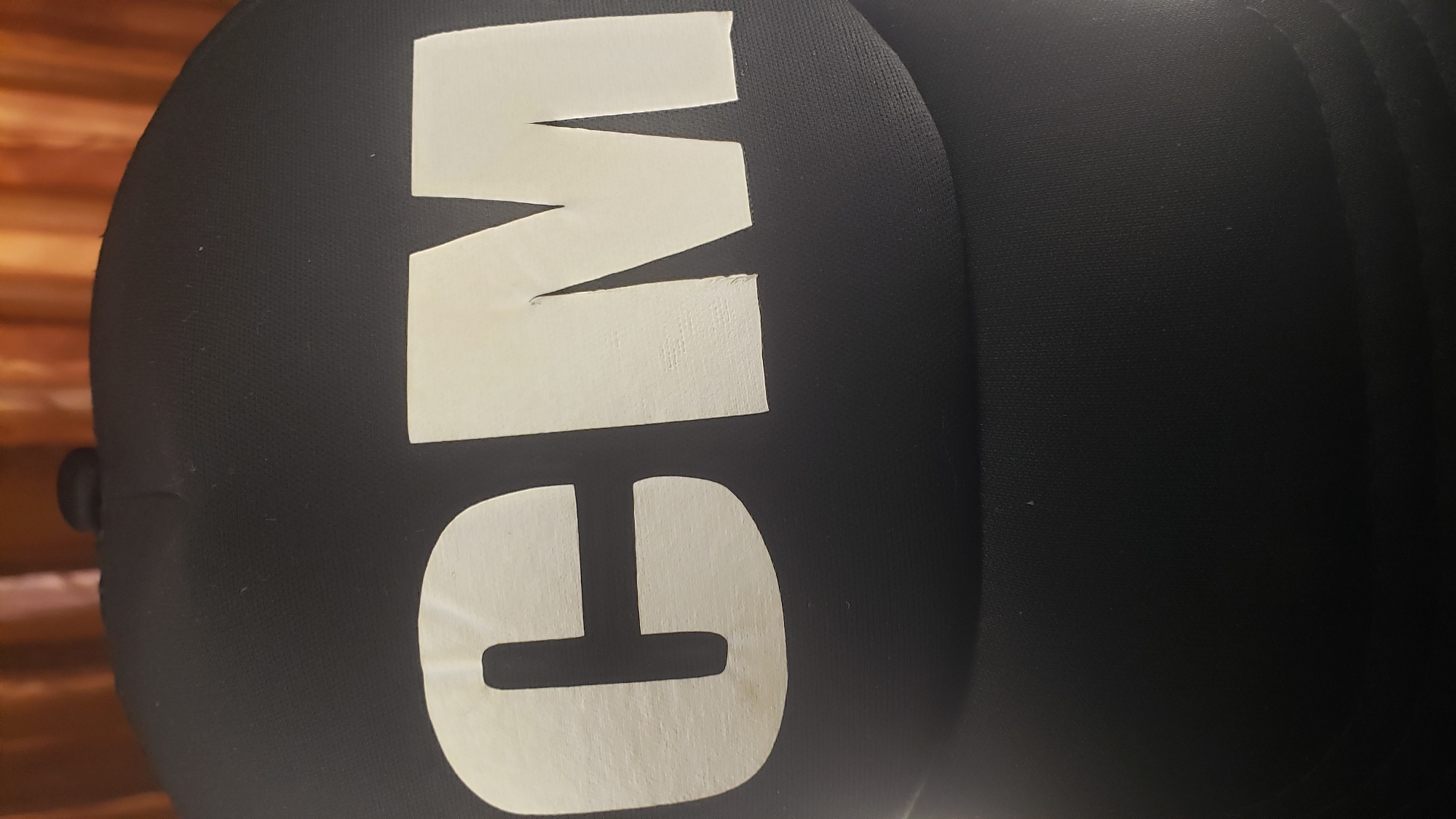 A close-up shot shows the letters "GM" applied to a black, rounded object. The letters appear to be made of white adhesive material. The background is a wooden wall.