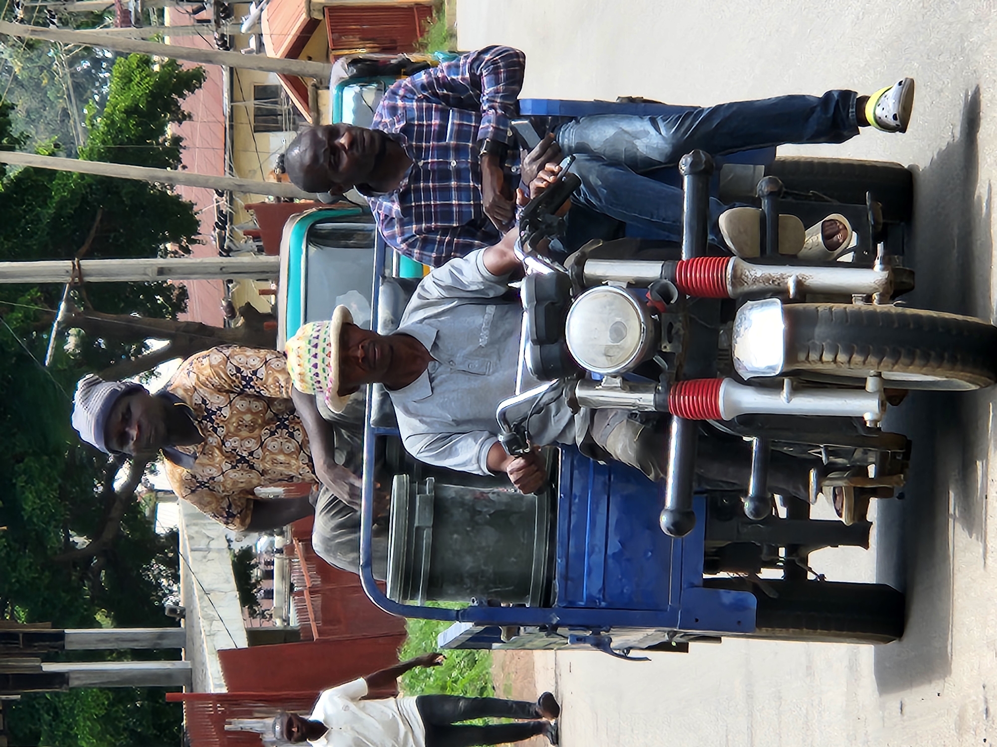 The image depicts three men in what appears to be a street scene. Two men are seated in a blue, motorized tricycle-like vehicle, while a third man stands to the left of the vehicle, facing away from the camera.
The man in the front seat of the vehicle is wearing a light gray shirt and a patterned hat. He is looking towards the left. The man in the back seat is wearing a blue and white checkered shirt and blue jeans. He is looking down at his hands, which are holding a smartphone. He is also wearing a watch on his left wrist. His right leg is extended, and he is wearing a shoe with a yellow band around the ankle.
The standing man is wearing a white shirt and dark pants. He is gesturing with his left arm.
The vehicle itself has a blue body and a metal frame. It has a round headlight on the front and appears to be a form of transportation. The background shows buildings with red roofs and trees, suggesting an outdoor setting. The lighting suggests it is daytime. There is no visible text in the image. The overall atmosphere seems casual, possibly a moment of pause during travel or a social interaction.