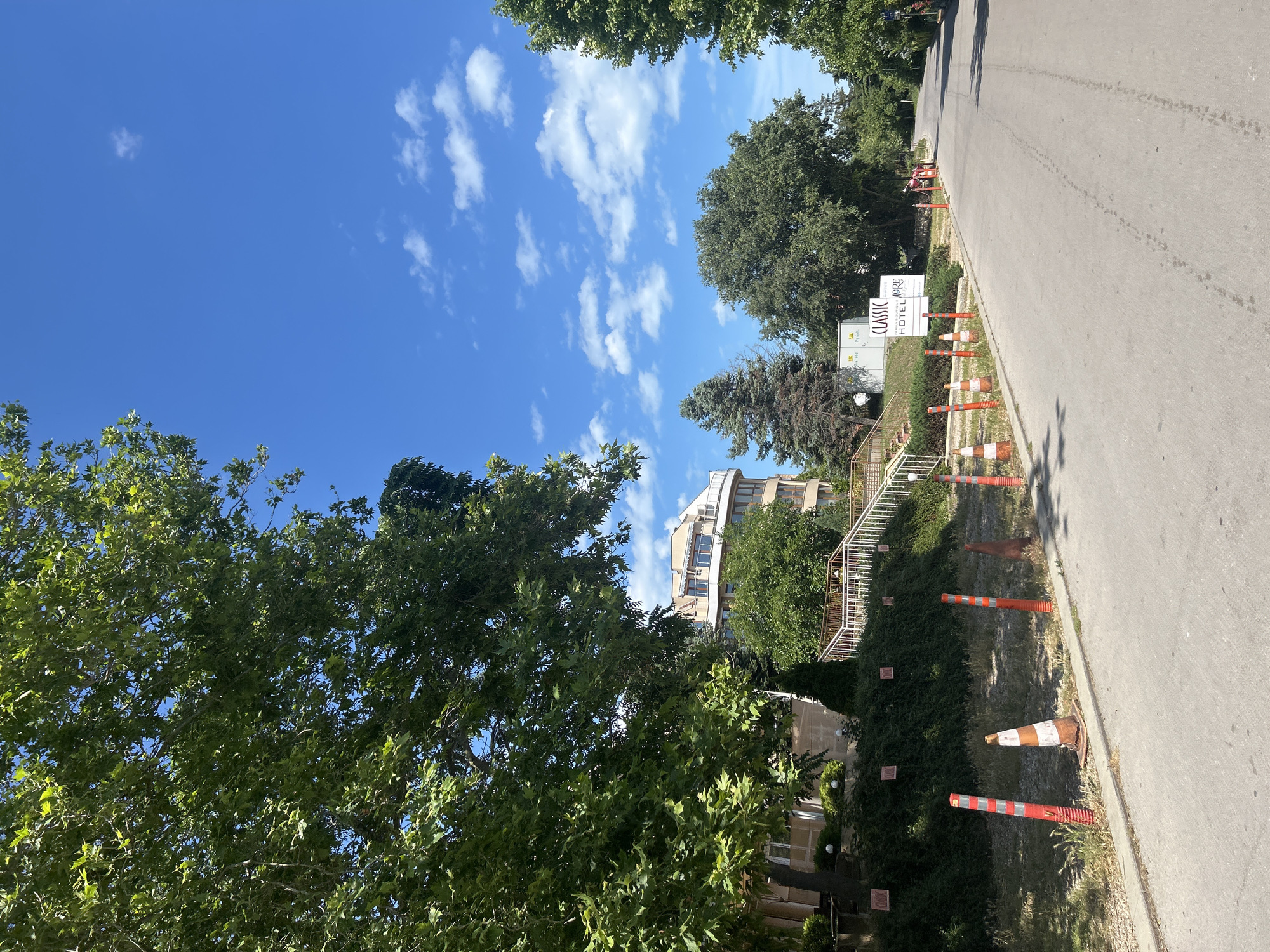 The image depicts a sunny day with a clear blue sky dotted with white clouds. The scene is an outdoor view of a paved road running alongside a green, grassy area bordered by a low hedge. Several orange and white traffic cones are lined up along the edge of the road, suggesting some form of temporary obstruction or caution.
In the background, a modern, multi-story building with curved balconies is visible, surrounded by lush green trees. To the left of the building, a sign is partially obscured by foliage, but the words "CLASSIC HOTEL" are clearly legible. The overall setting appears to be a well-maintained urban or suburban environment, possibly near a hotel or residential area. The bright sunlight and clear sky indicate it is daytime, likely late morning or early afternoon. No people or animals are visible in the frame. The mood is serene and peaceful, characteristic of a pleasant day.
