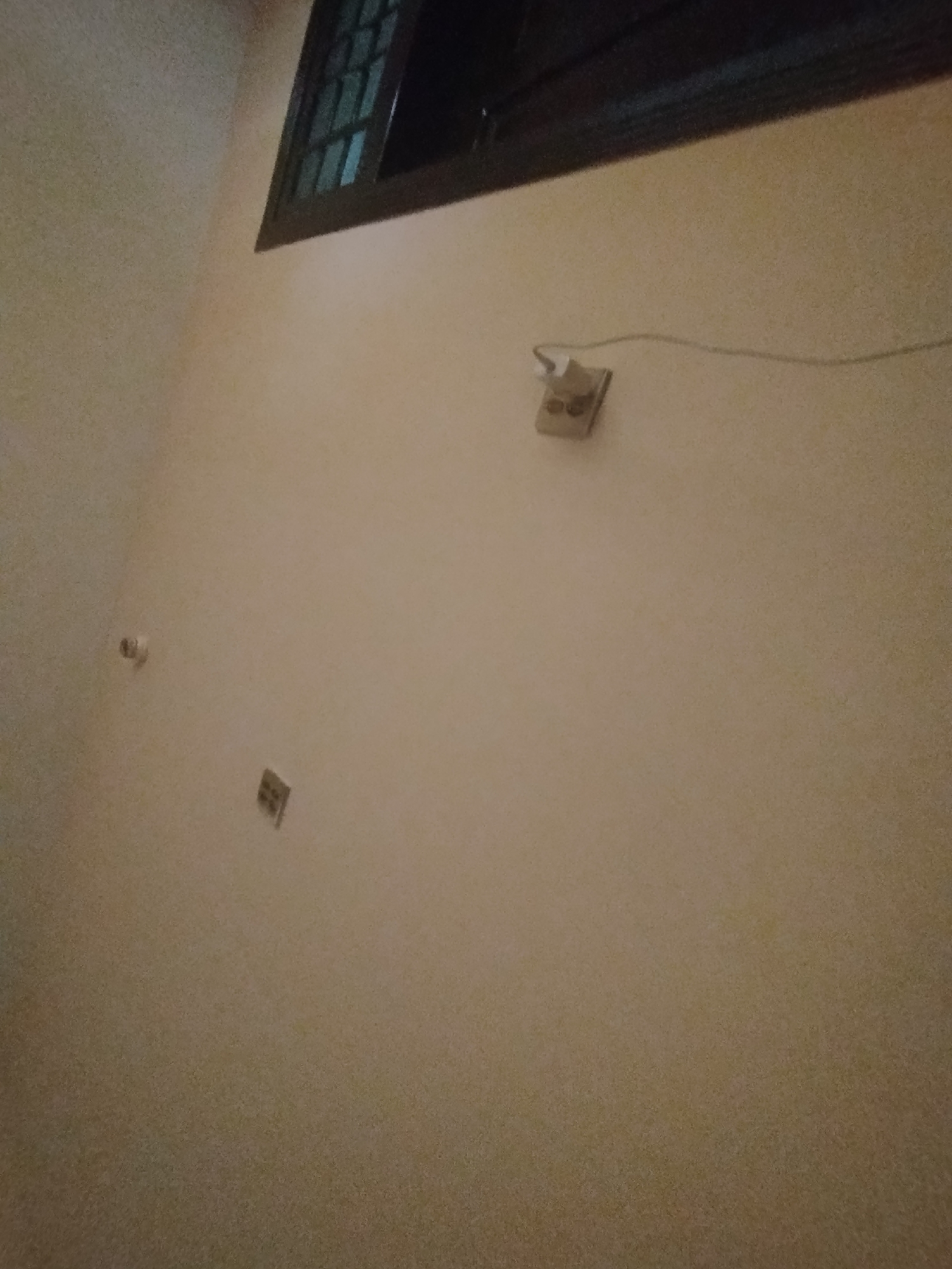 The image depicts an interior scene, likely a room with light-colored walls. Near the top of the image, a dark-colored window frame with a patterned glass design is visible. Mounted on the wall are two electrical outlets. One outlet, located in the upper right portion of the image, is a duplex outlet with a light-colored casing. A white electrical cord extends from this outlet. A second, smaller square object, possibly another outlet or a light switch, is located on the wall in the lower left quadrant. A third small, rectangular object, also possibly an electrical component, is situated on the wall below the upper outlet. The lighting in the room appears to be dim. There are no people or overt actions depicted in the image. The environment suggests a residential or office setting within the city of Lassan, Pakistan.