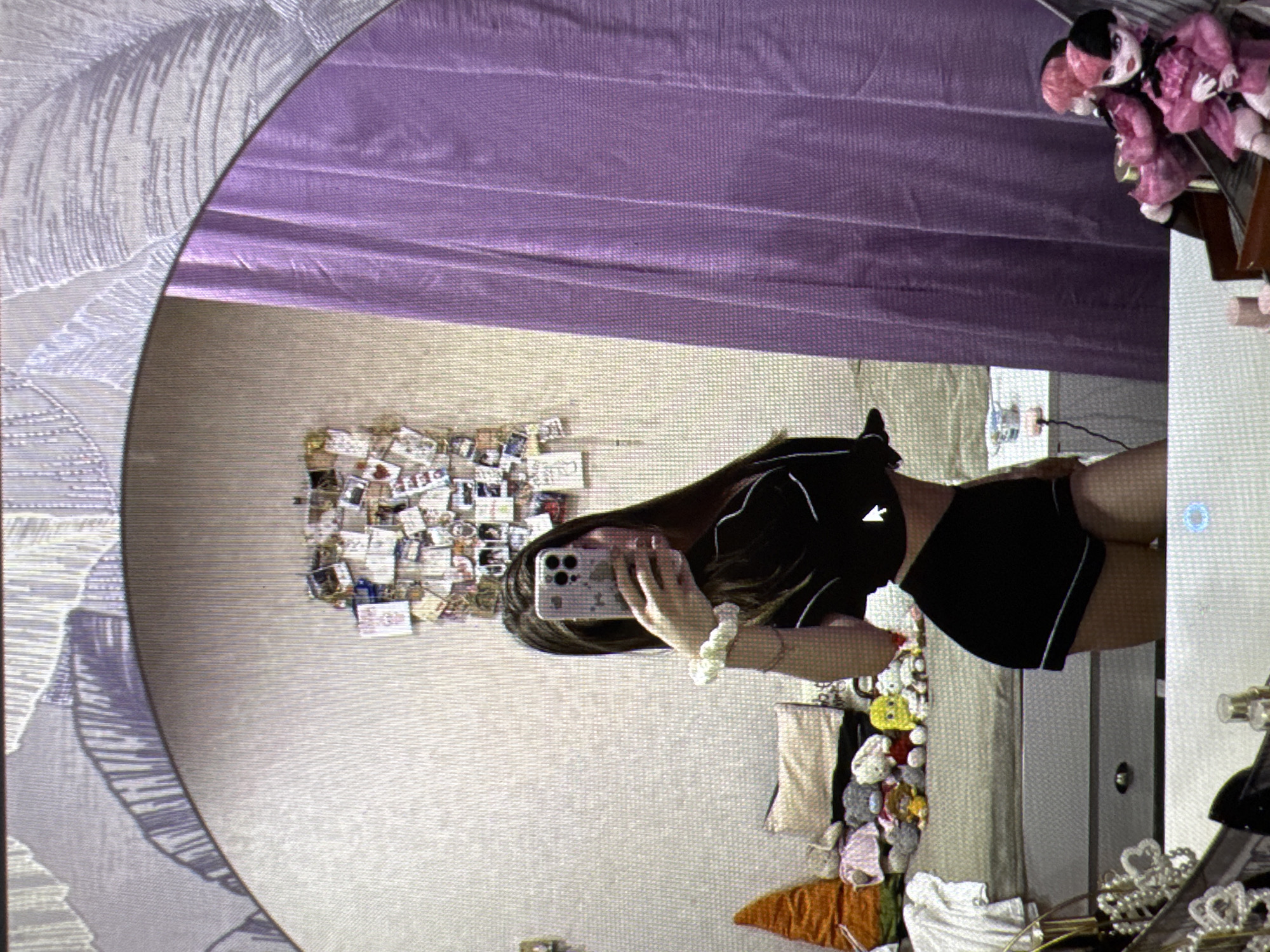 The image is a selfie taken in a room. A young woman with long brown hair is reflected in a large oval mirror. She is wearing a black cropped top and black shorts with white piping. She is holding a smartphone in her right hand, which is angled to capture her reflection. A scrunchie is visible on her left wrist.
Behind the woman, the room is visible. The wall behind her has a collage of photos and papers pinned to it. To her right, a purple curtain hangs. In the foreground, the reflection shows a bed with several stuffed animals and an orange conical object. Below the bed, a chest of drawers is visible. In the lower right corner, two dolls with pink and black hair are seated on what appears to be a wooden shelf or display.
The scene is domestic, likely a bedroom. There is no indication of the location being in the city of Castelo Branco, Portugal.