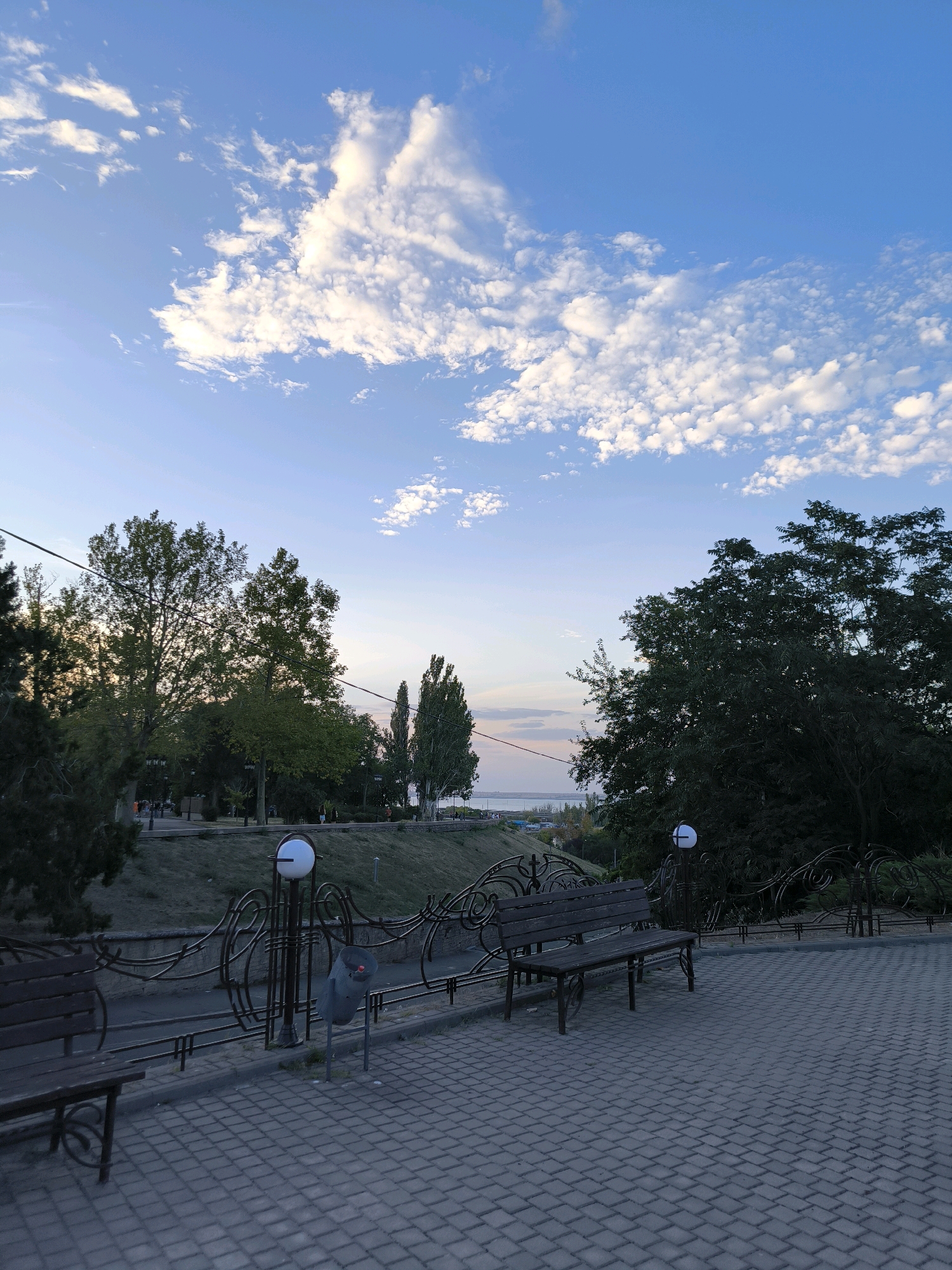 The image depicts a park area with a paved walkway in the foreground, flanked by decorative metal fencing. Two wooden benches are visible, facing towards a grassy slope and trees. Beyond the trees, a body of water, likely the Southern Bug River, can be seen in the distance, under a partially cloudy blue sky. The overall scene suggests a tranquil, early evening setting in a public park. This location is characteristic of the waterfront promenade in Mykolaiv, Ukraine, a popular spot for residents and visitors.