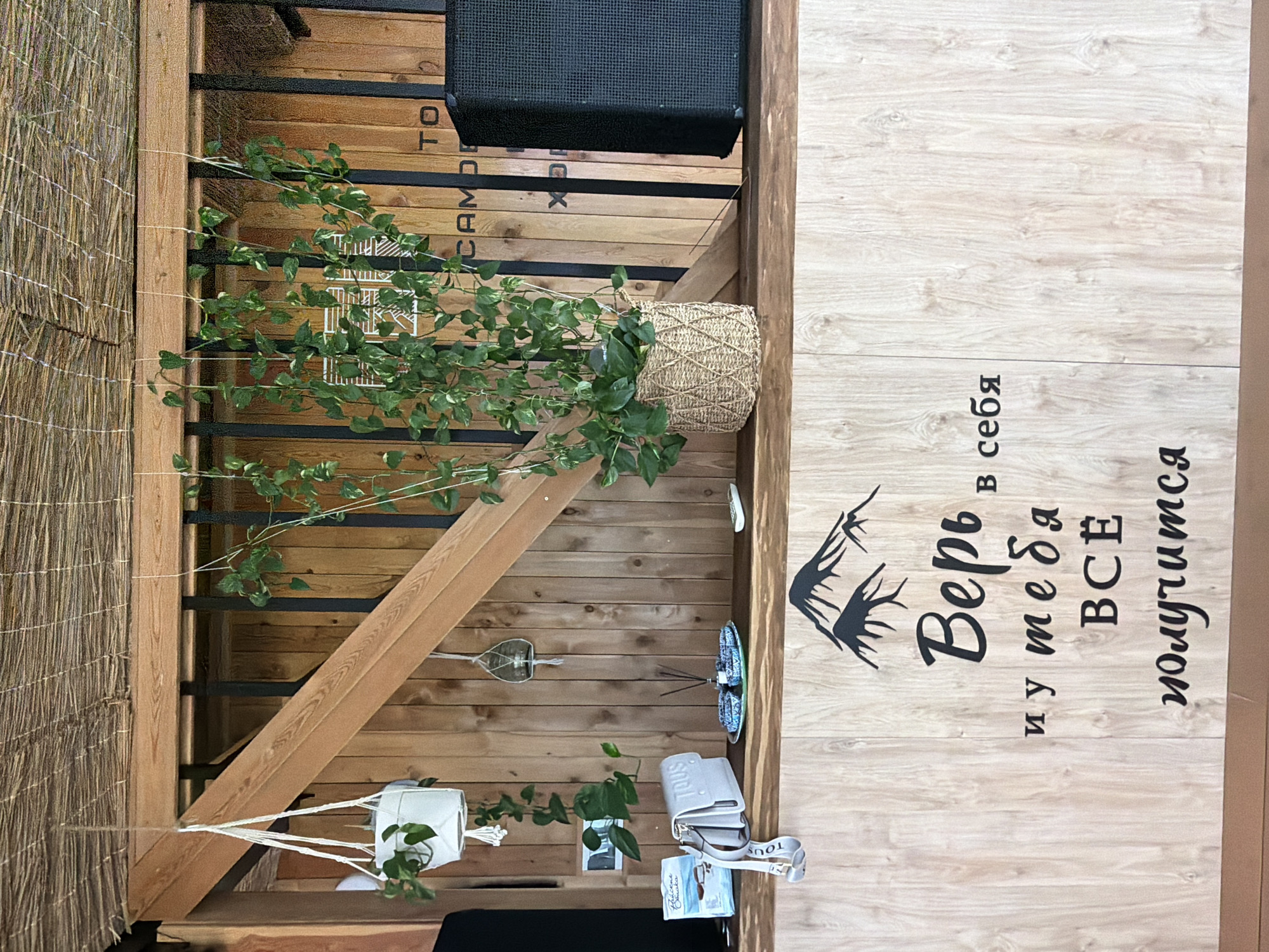 A medium shot captures a wooden bar counter with a patterned wooden wall behind it. A basket-style planter with a large green vine plant is placed on the counter. To the right of the plant, a black speaker is visible. Above the counter, the wooden wall is adorned with text that appears to be Russian and English branding, with the partial word "CAMDE" visible. To the left of the plant, a light-colored purse with the brand name "TOUS" is placed on the counter, next to a small package and a candle-holder hanging from a string. Below the counter, the wooden surface is decorated with a black silhouette of mountains and Russian text. The overall scene appears to be an indoor or semi-outdoor setting, possibly a cafe or bar. The location is specified as Nizhnyaya Shilovka, Russia.