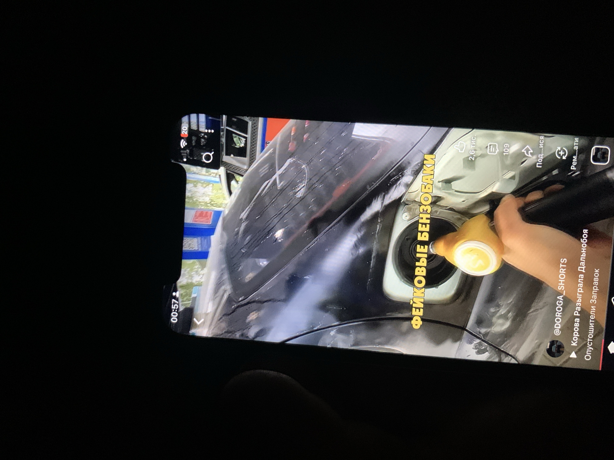 The image shows a smartphone screen displaying a short video. The video appears to be taken at a gas station, with a close-up shot of a hand holding a yellow gas pump nozzle inserted into a car's fuel filler neck. The car is black and appears to be wet. Text overlaid on the video reads "ФЕЙКОВЫЕ БЕНЗОБАКИ" (Fake Fuel Tanks) in yellow lettering. Below this, a YouTube channel name "@DOROGA_SHORTS" is visible, followed by two video titles: "Корова Разыграла Дальнобоя" (Cow Tricked a Trucker) and "Опустошители Заправок" (Gas Station Emptiers). On the right side of the screen, social media interaction icons like "likes" (2.6k), "comments" (109), and "share" are displayed. The top of the phone screen shows the time "00:57" and signal icons. The background is dark, indicating the photo was taken in low light conditions. The image does not contain any information or visual cues to indicate the location is the city of Zhmerynka, Ukraine.