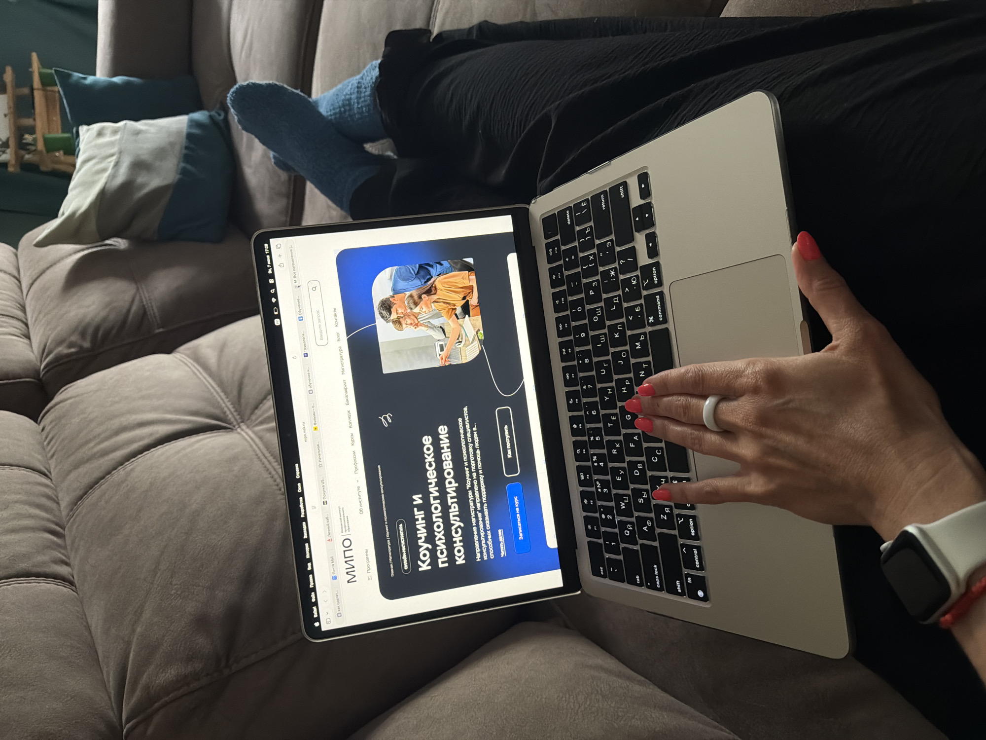 In an indoor setting in Birobidzhan, Russia, an unidentified individual is depicted reclining on a light brown, tufted upholstered sofa. The person's lower torso is covered by a textured black garment, and their feet, crossed at the ankles, are clad in blue knitted socks. A silver Apple MacBook laptop is positioned on their lap, with its screen displaying the website "mipo.msk.ru" in the Safari browser. The website content is in Russian, featuring the title "МИПО" (Moscow Institute of Professional Education) and a prominent section on "Коучинг и психологическое консультирование" (Coaching and Psychological Counseling), with options to "Записаться на курс" (Enroll in the course) and "Как поступить" (How to apply). An image of four individuals looking at a computer screen is part of the website's design. The laptop's macOS interface shows the date as "June 7" and the time as "17:29." The laptop keyboard features both Latin (QWERTY) and Cyrillic characters. The person's right hand, adorned with red nail polish and a white ring on the ring finger, is poised over the laptop's trackpad and keyboard area. A white Apple Watch and a red string bracelet are visible on the person's left wrist. In the background, two decorative pillows are visible on the sofa: one in shades of light blue, grey, and white, and another solid teal. A dark teal-colored wall and a partially obscured wooden shelf are visible behind the sofa.