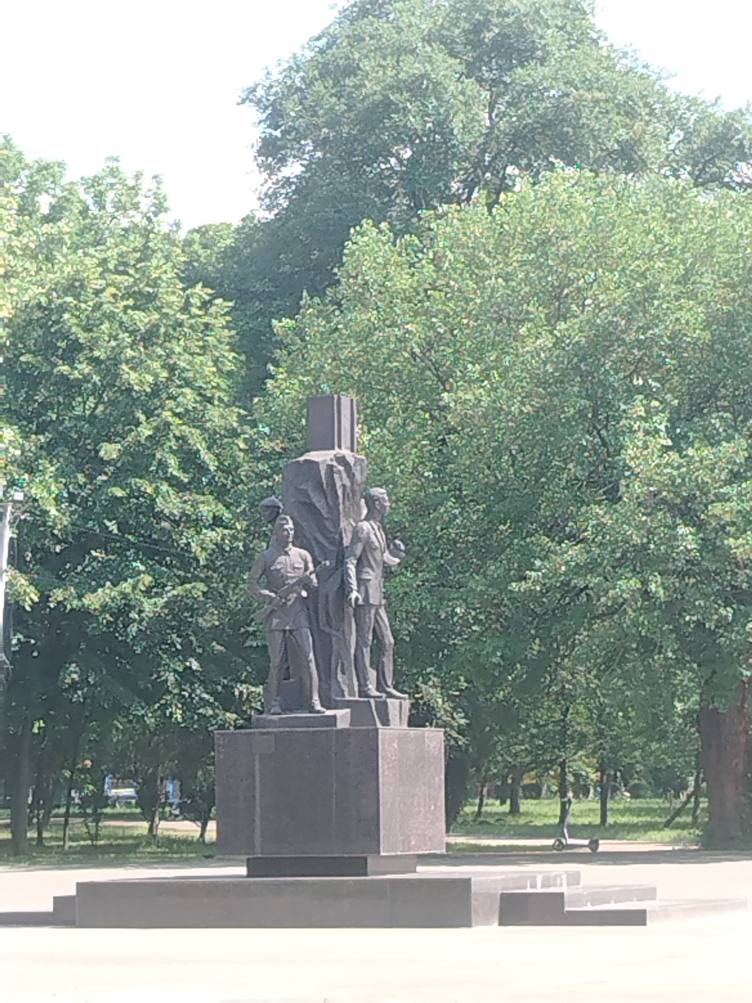 This is a medium shot photograph of a monument in a park setting. The monument is made of dark stone and depicts three figures standing on a raised base. The figures are in military attire, with one figure in the foreground holding a rifle. Behind the monument, there are lush green trees with dense foliage, indicating a warm, sunny day. In the background, a person is seen riding a scooter on a paved path. The monument is likely a memorial, possibly related to military history, and is situated in a public park in Krasnodar, Russia. The overall scene suggests a peaceful, everyday moment in an urban green space.