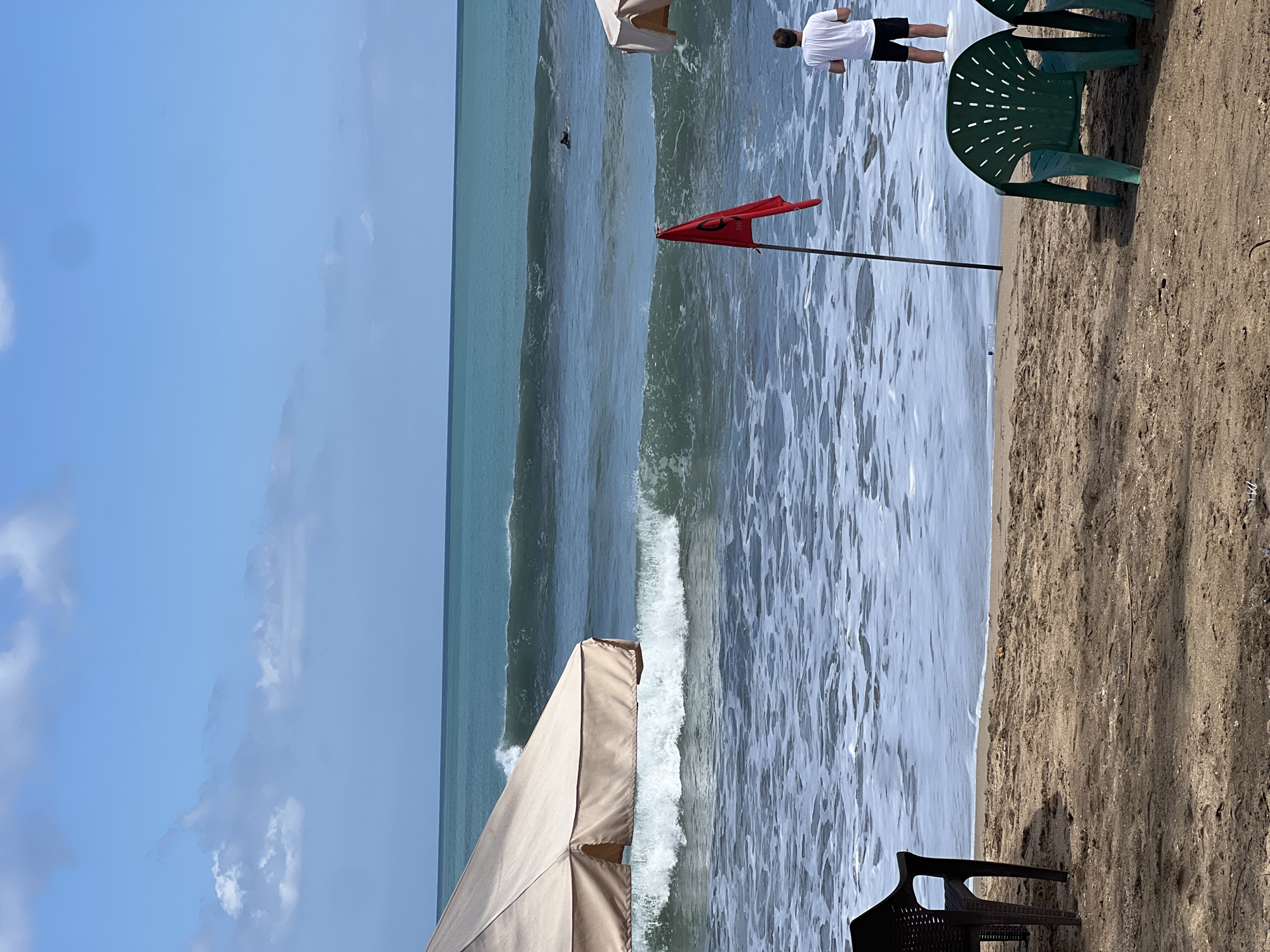 The image depicts a beach scene in Kuta, Indonesia, on a sunny day with a partly cloudy sky. In the foreground, two plastic chairs, one dark brown and one green, sit on the sandy beach. A large, beige beach umbrella is partially visible on the left side of the frame.
In the middle ground, the ocean displays moderate waves with white foam. A red flag, likely indicating swimming conditions or a safety warning, is planted in the shallow water. A person wearing a white t-shirt and black shorts stands in the water, facing away from the camera, near the red flag. Further out in the ocean, a surfer is visible riding a wave.
The background shows the vast expanse of the turquoise ocean meeting the horizon under a blue sky with scattered white clouds. The overall impression is a typical beach day in Kuta, with elements of leisure and water sports present.