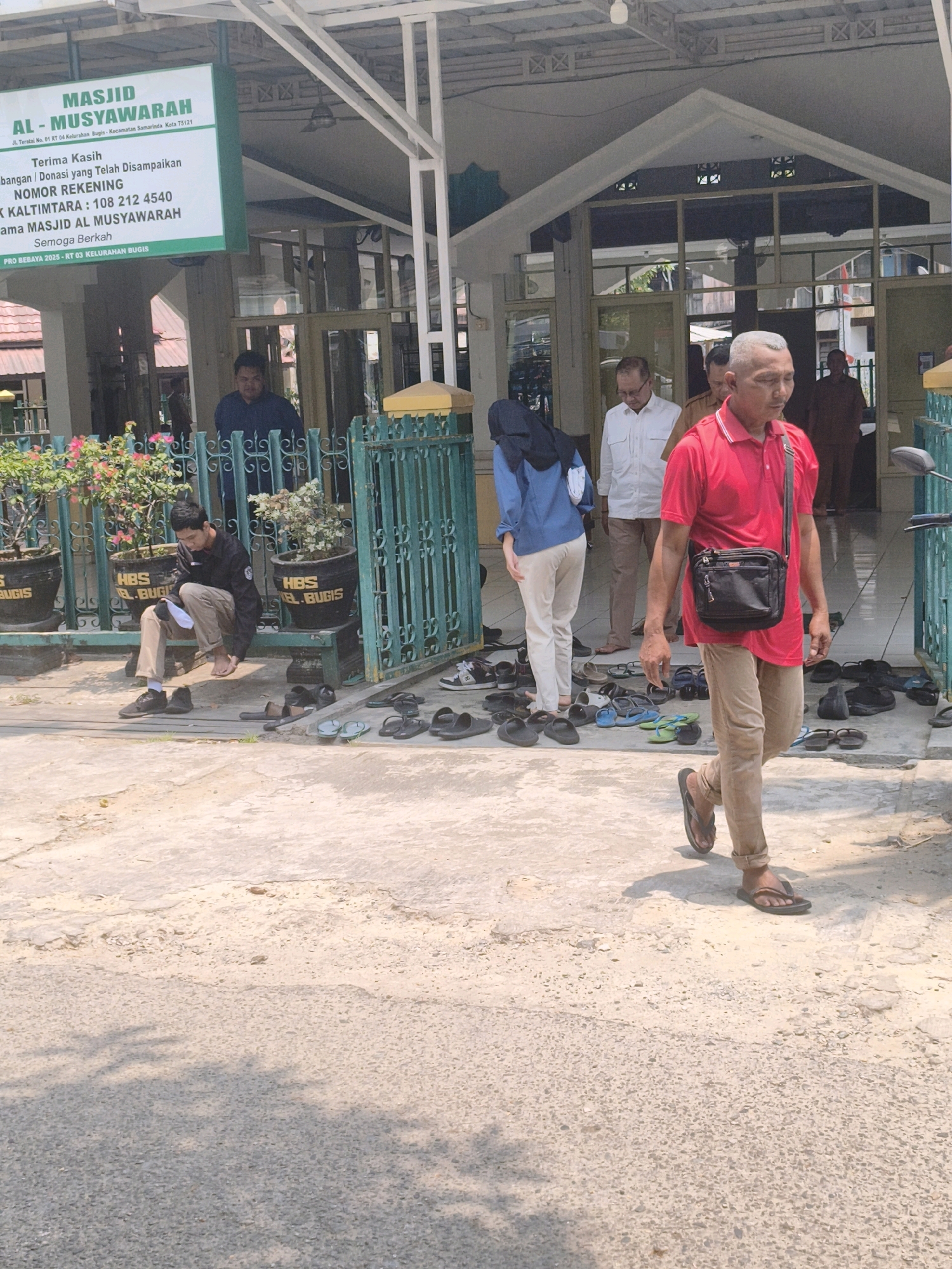The image depicts the entrance of the AL-MUSYAWARAH Mosque, located at Jl. Teratai No. 01 RT 04 Kelurahan Bugis, Kecamatan Samarinda, Kota 75121, in Samarinda, Indonesia. A man in a red polo shirt and tan pants walks away from the mosque entrance, carrying a black crossbody bag. To his left, a person wearing a blue long-sleeved shirt and light-colored pants, with a dark hijab, stands near the entrance. Further left, a man in a dark jacket and tan pants sits on a bench, looking down at something in his hands. Several potted plants with pink and white flowers are visible near the entrance, with one plant labeled "HBS BUGIS." Various pairs of sandals and flip-flops are scattered on the ground near the mosque entrance. The mosque features a green metal fence and a peaked roof structure. The ground outside the mosque is a paved, rough surface. In the background, other individuals can be seen near the mosque.