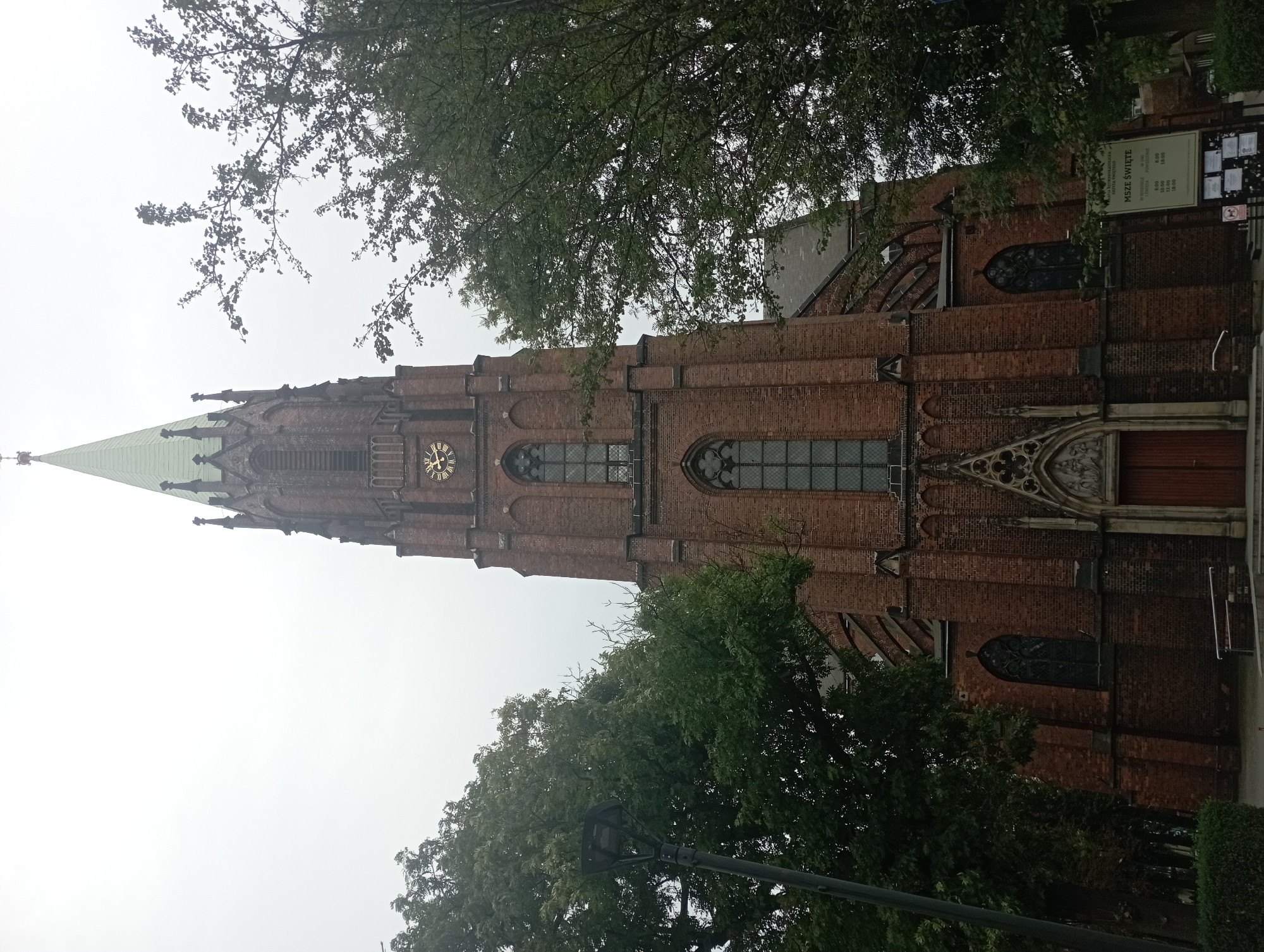 The image shows a tall, brick church with a green steeple against a hazy, overcast sky. The church has Gothic architectural elements, including pointed arches over the windows and entrance. A clock face with Roman numerals is visible on the tower. Trees with dense green foliage flank the church on either side, partially obscuring the view of the building's wings. A lamppost stands to the left of the church. A sign with Polish text, including "MSZE ŚWIĘTE" (Holy Masses), is posted to the right of the entrance. The foreground shows a paved area with some steps leading up to the church entrance, and a bench to the left. The overall scene suggests a slightly damp or humid day in an urban environment. This is the Holy Cross Church (Kościół Świętego Krzyża) in Siemianowice Śląskie, Poland.