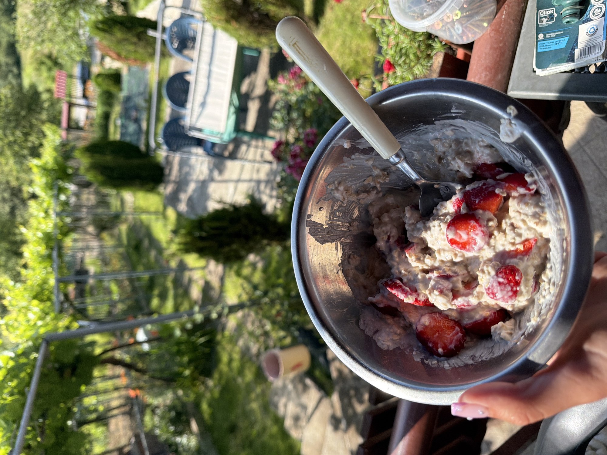 A person is holding a metal bowl of oatmeal with strawberries. The bowl is in the foreground, with a spoon inside. In the background, there is a garden with green trees, plants, and outdoor furniture. The location is Balinovtsi, Bulgaria. The image appears to be taken outdoors on a sunny day.