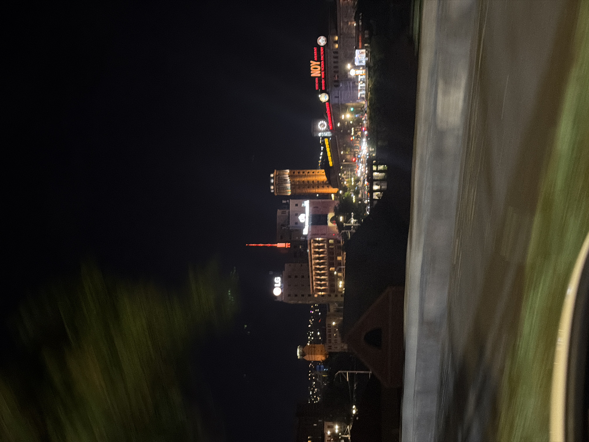 This is a nighttime photograph taken from a moving vehicle, likely a car, overlooking a city illuminated by artificial lights. The perspective suggests the viewer is on an elevated road or overpass. In the foreground, to the right, is a concrete barrier and blurred green foliage, indicating movement and a roadside setting.
The main subjects of the image are buildings in the cityscape. These structures vary in height and architectural style, with some appearing modern and others older. Many of these buildings are lit from within and by external signage, creating a vibrant urban glow against the dark night sky. One prominent building features a large, circular "LG" logo, indicating a recognizable brand presence. Another building has a tall, red, spire-like structure on top, illuminated by red lights. The overall scene is one of an active city at night, with a bustling atmosphere suggested by the density of lights and buildings.
Visible text includes the "LG" logo and "NOY" in red neon lettering, which appears to be part of a larger sign on one of the buildings. There are also other, less legible signs and illuminated advertisements. The time of day is clearly night, and the weather appears clear as the city lights are sharp and visible. The location is suggested to be Yerevan, Armenia, which is supported by the presence of the "NOY" brandy distillery sign, a well-known Armenian brand. The blurry nature of the foliage and the road suggests the photograph was taken while in motion, possibly from a highway overlooking the city. The overall mood conveyed is one of urban energy and nighttime activity.