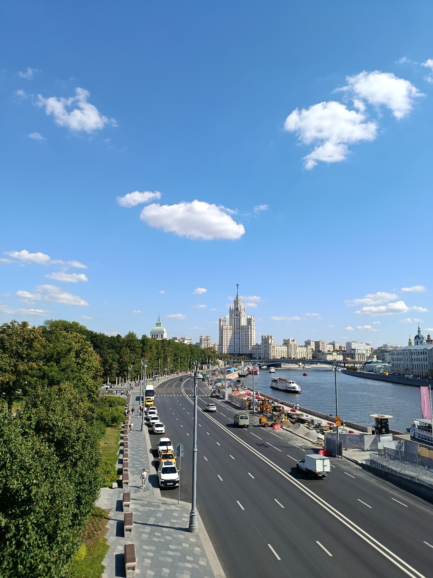 An eye-level, full shot captures a bright, sunny day in a city with a large river flowing through it. The sky is a clear, vibrant blue, dotted with fluffy white clouds. On the left side of the frame, a dense line of lush green trees borders a pedestrian walkway. The walkway itself is paved with gray and white patterned tiles and features a row of modern, light-colored benches. Several people are walking along the walkway.
Adjacent to the walkway is a multi-lane road carrying a steady flow of traffic. Various vehicles, including white cars, yellow taxis, and a white truck, are visible. The road is marked with double white lines.
On the right side of the road, the river dominates the landscape. Several boats, including a large white passenger boat, are navigating the water. Construction is underway along the riverbank, with barriers, piles of gravel, and construction equipment visible. Beyond the river, the city skyline is characterized by a mix of architectural styles, most notably a tall, ornate white building with a spire, likely a prominent landmark, and other buildings in various shades of white and beige. The scene is active, with transportation and construction occurring under a clear sky.