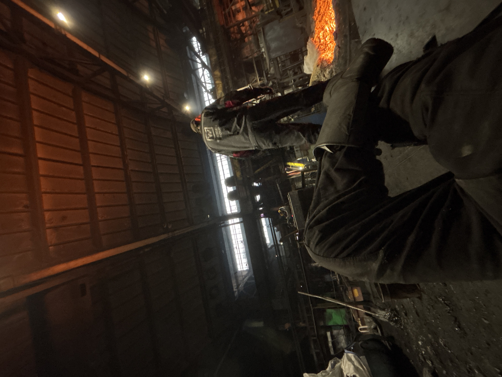 The image depicts an interior industrial setting, possibly a foundry or metalworking facility, with a low-angle perspective showing a person's legs and feet in the foreground. The individual is wearing dark work pants and appears to be sitting or resting. In the midground, a person in a dark jumpsuit with a white logo on the back is standing, facing away from the camera. They are positioned near a bright, glowing orange material on the ground, which resembles molten metal or a heat source. The environment appears gritty and industrial, with visible tools, debris on the floor, and large machinery or structures in the background. The ceiling is high and industrial, with visible beams and lighting. The overall lighting is dim, with the brightest illumination coming from the glowing material and some overhead lights. No specific landmarks within Homyel, Belarus, are identifiable from the visual information provided.