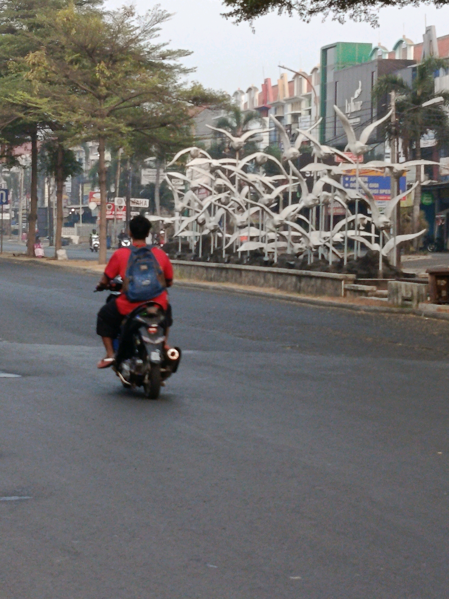 In Bekasi, Indonesia, a person wearing a red shirt and a blue backpack rides a motorcycle on a paved road. The rider is viewed from behind, and the motorcycle is black with a silver exhaust pipe. The road is bordered by a low stone wall with some vegetation. Behind the wall, a large sculpture of white birds in flight is visible, with a modern building complex in the background. Trees line the left side of the road, with some buildings and street signs visible in the distance. The sky is overcast.