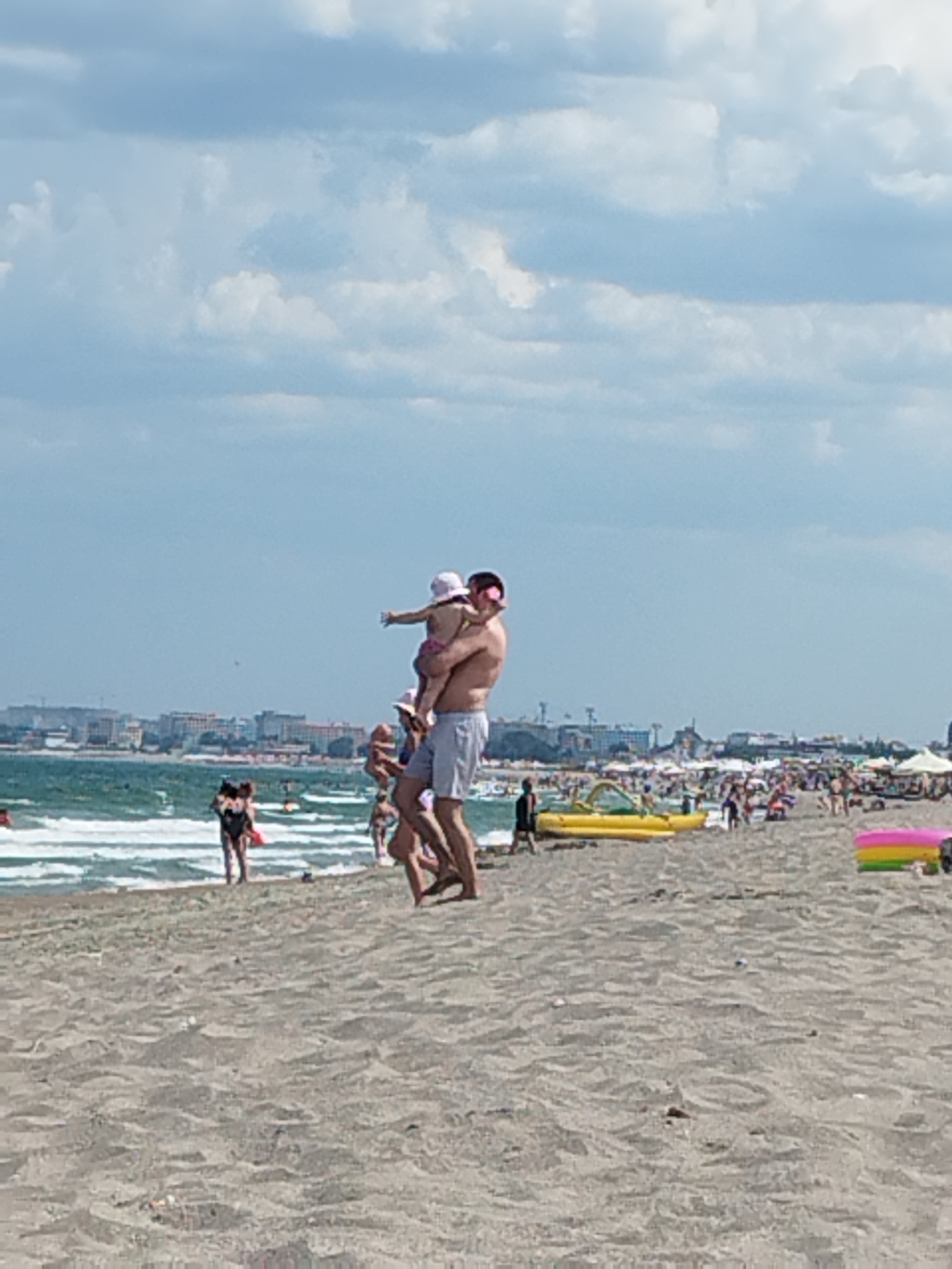 The image depicts a crowded beach scene in Constanța, Romania, under a partly cloudy sky. In the foreground, a man is carrying a young child on his shoulders, with the child reaching out an arm. Other beachgoers are visible in the middle ground, walking on the sand and swimming in the sea. The background shows a cityscape with numerous buildings and a long stretch of coastline. Various beach accessories, such as umbrellas, lounge chairs, and inflatable toys, are scattered across the sand. The overall atmosphere suggests a lively and bustling summer day at the beach.