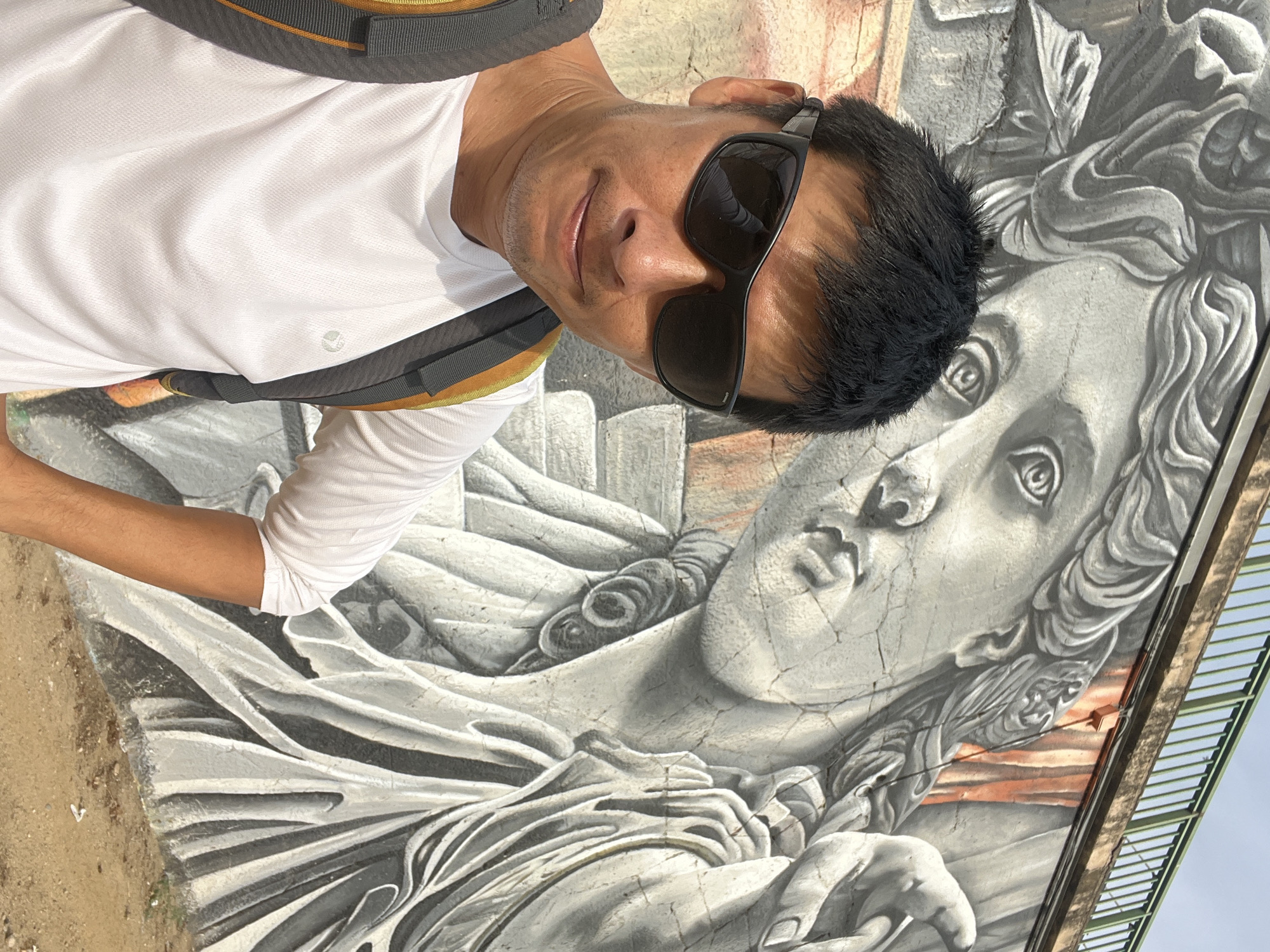 A man wearing sunglasses and a white t-shirt is taking a selfie in front of a mural on a wall. The mural depicts a large, grey, classical-style statue with flowing hair and draped clothing. The man is positioned in the lower left corner of the image, with his face and upper body visible. He has dark hair and is wearing a backpack with grey and yellow straps. The background is a textured wall with the large mural. The ground in front of the wall appears to be dirt and gravel. A green metal railing is visible at the top of the image.
This mural is located on the wall of the Balcó del Mediterrani in Tarragona, Spain. The Balcó del Mediterrani is a viewpoint overlooking the Mediterranean Sea, and the murals on its walls often depict historical or mythological themes.