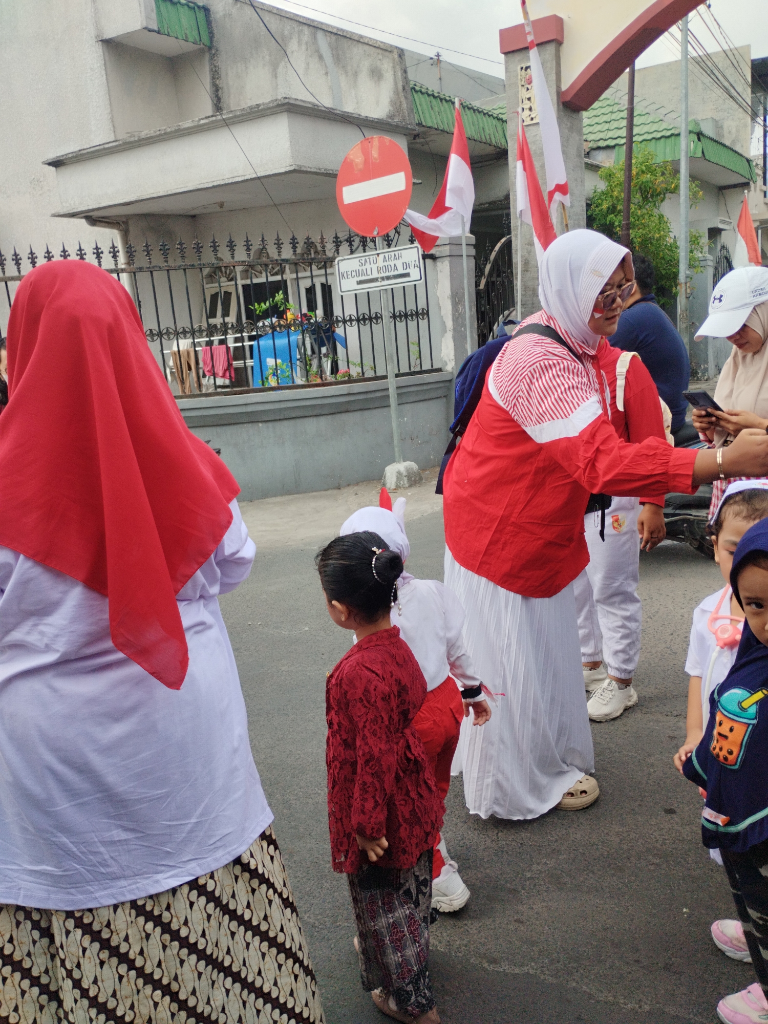 The image shows a street scene in Surabaya, Indonesia, likely during a celebration, given the presence of Indonesian flags and people dressed in red and white attire. In the foreground, a woman wearing a red and white striped jacket and white skirt is interacting with her phone, with a child partially visible in front of her. To the left, a person is seen from the back, wearing a red hijab and a white top. Several other individuals, including children, are scattered throughout the scene, some in festive clothing. A red and white Indonesian flag is visible on a pole in the background, as well as a red circular sign with a white horizontal bar. The environment appears to be a residential street with a house and a fence visible.
