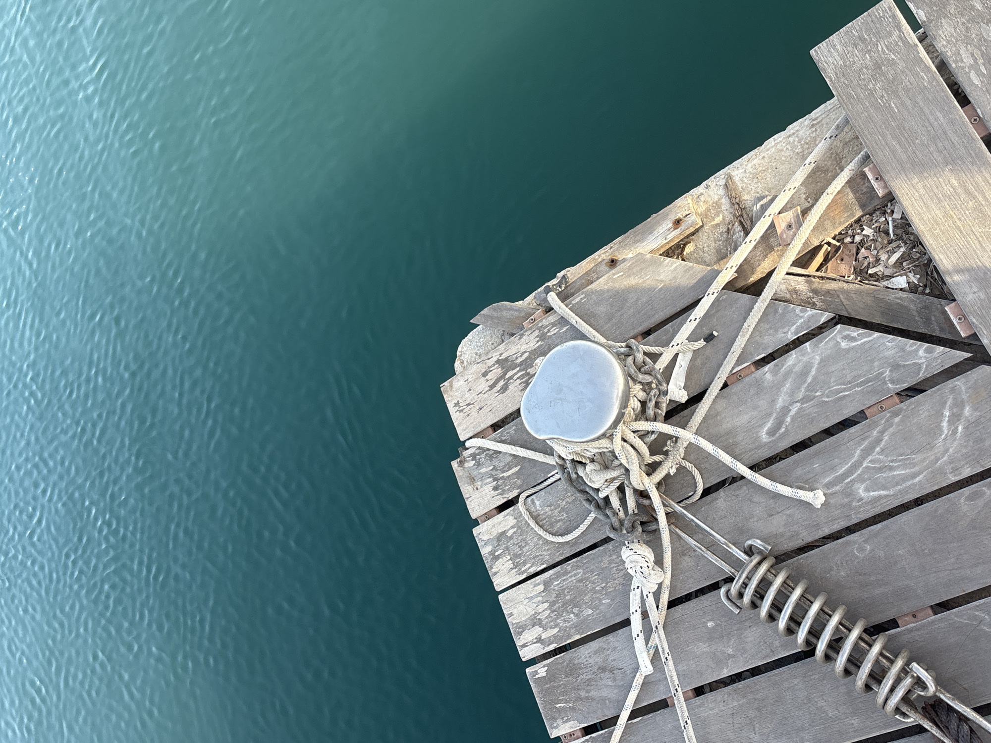 The image shows a close-up view of a weathered wooden dock with ropes and a metal cleat, beside a body of teal-colored water. The dock planks are oriented diagonally, and some show signs of wear and tear, with gaps filled with debris. Several white ropes are coiled and attached to the cleat, and one rope extends towards the lower left, attached to a metal spring. The water is calm with a rippled surface, reflecting the light. The scene appears to be from Urbanización Cabo Roig, Spain, a coastal area known for its marinas and waterfronts. There are no people or animals visible in the frame.