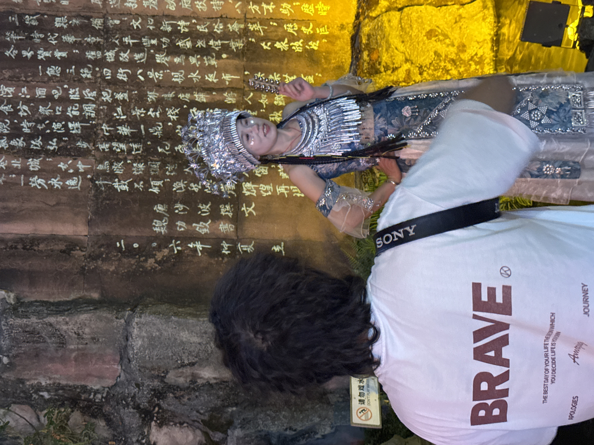 The image depicts a nighttime scene in Yuzhong District, China, with a person in traditional attire posing against a stone wall with engraved text. The person, a woman, wears an elaborate headdress and ornate silver jewelry, including a large necklace and armbands. Her dress features intricate blue and white patterns and embellishments. She holds a small, bell-like object in her right hand. In the foreground, partially obscuring the woman, is the back of another person wearing a white t-shirt with the word "BRAVE" printed in large, dark red letters. A black camera strap with the "SONY" logo is visible across the t-shirt. The stone wall behind them is illuminated with a warm yellow light, highlighting the white-colored Chinese characters carved into its surface. A small sign with red text and an image of a person playing is visible on the left side of the image, with the words "DON'T PLAY WATER" in English below it. The overall environment suggests a cultural or tourist location.