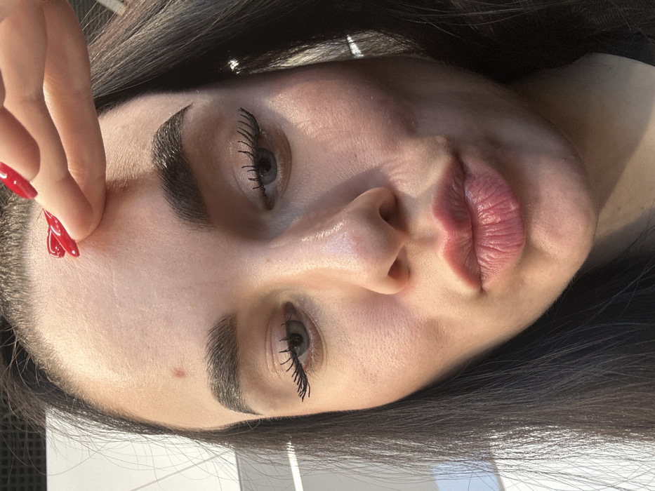 The image displays a close-up, frontal portrait of an adult female. Her dark hair is visible on the sides of her face. She has light-colored eyes, directed downwards, with prominent, dark eyelashes, indicative of mascara use. Her eyebrows are thick and dark. Her lips are full, pinkish in color, and pursed into a pout. A small, reddish raised lesion or blemish is present on her forehead, located to the left of the midline. A right hand, with fingers exhibiting bright red nail polish, is shown touching or gently pinching the skin on her upper forehead. The background is out of focus, consisting of indistinct white and grey shapes, consistent with an indoor setting under artificial light. The image contains no visual information to establish its location within the city of Saint Petersburg, Russia, or any specific geographic area.