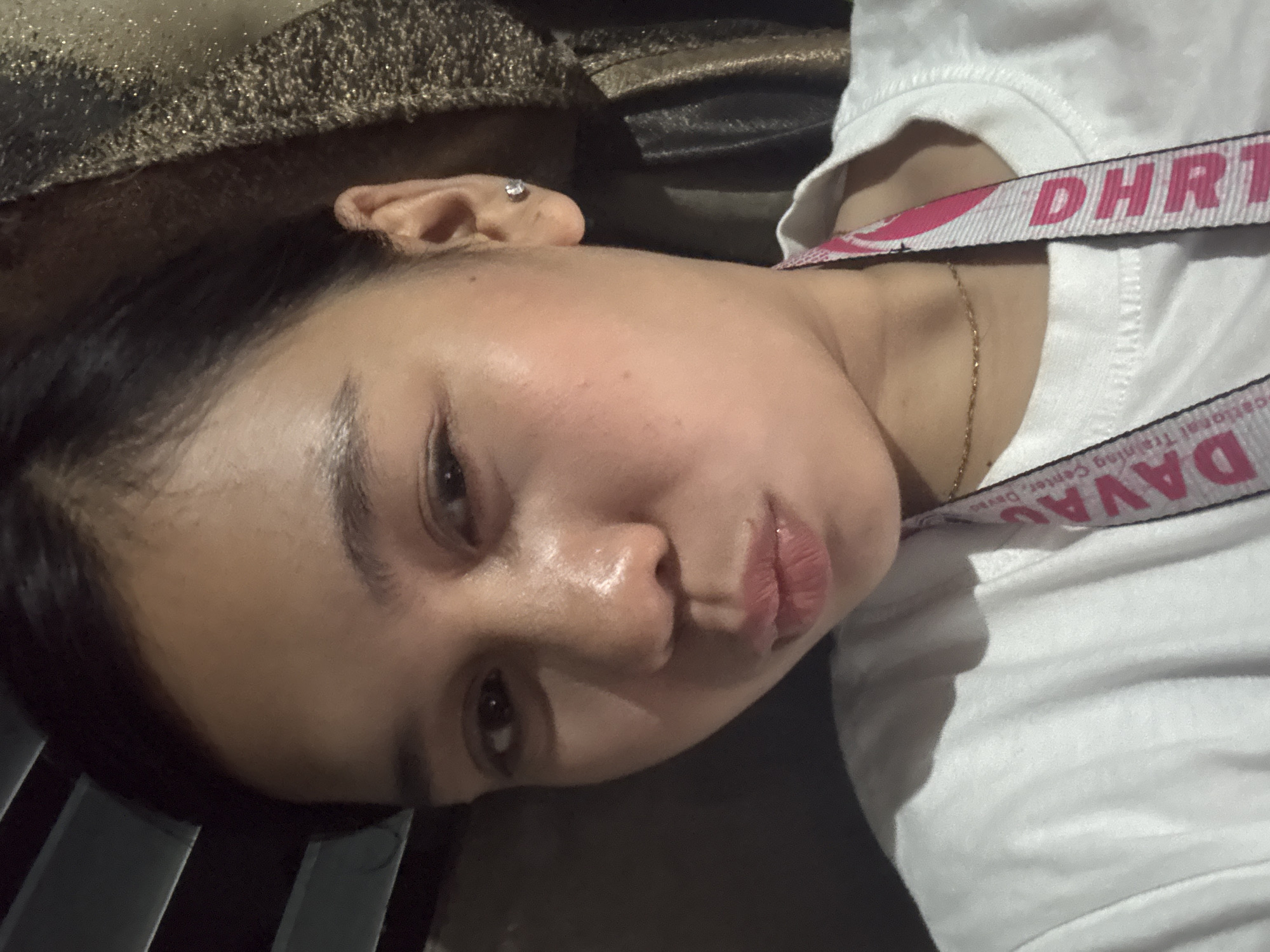 The image is a close-up selfie of a young woman, likely taken indoors. She has dark hair pulled back, and is wearing a white t-shirt. A lanyard with the text "DAVAO" and "DHRI" is visible around her neck, suggesting a possible connection to Davao City, Philippines, or an organization with a presence there. The background is dark and out of focus, with some hints of blinds on the left and a textured fabric on the right. The lighting suggests it is not daytime. The woman's face is the primary focus, with her lips puckered.
