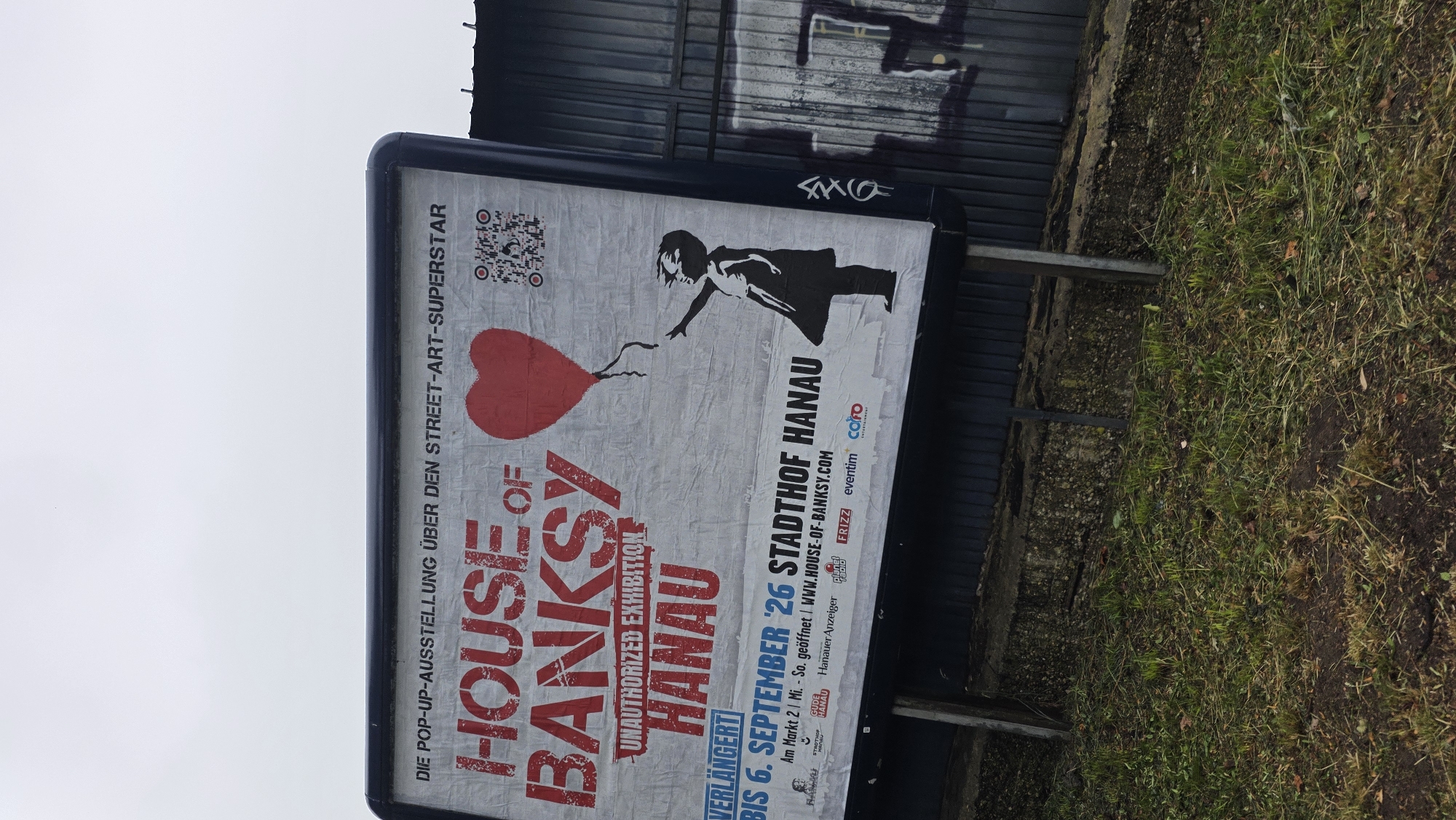 A vertical, eye-level shot captures a billboard advertising an unauthorized Banksy exhibition in Hanau. The billboard features the title "HOUSE OF BANKSY" in large red stencil letters, with the subtitle "UNAUTHORIZED EXHIBITION HANAU" below it. To the right of the text, a red balloon in the shape of a heart is depicted, with a QR code and a silhouette of a girl holding the balloon's string to its left. The bottom portion of the billboard indicates that the exhibition is extended until September 6, '26, and provides location details: "STADTHOF HANAU" along with website and sponsor information. The billboard is mounted on a metal frame and stands against a dark, corrugated metal fence. The foreground is a grassy area with some dry leaves and patchy soil. The sky is overcast and pale.