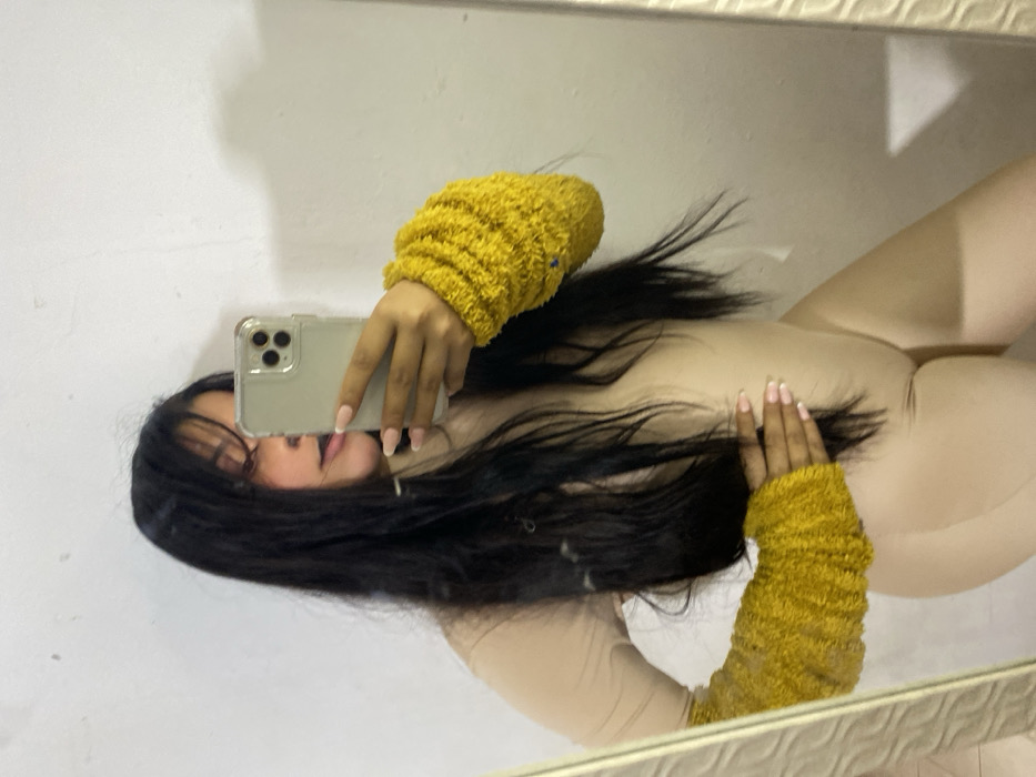 A selfie in a mirror shows a woman with long, dark hair. She is holding a smartphone in front of her face, angled slightly downward. Her nails are painted with a white tip. She is wearing form-fitting beige clothing and mustard-colored, textured hand warmers or sleeves. The background is a blurred white wall.