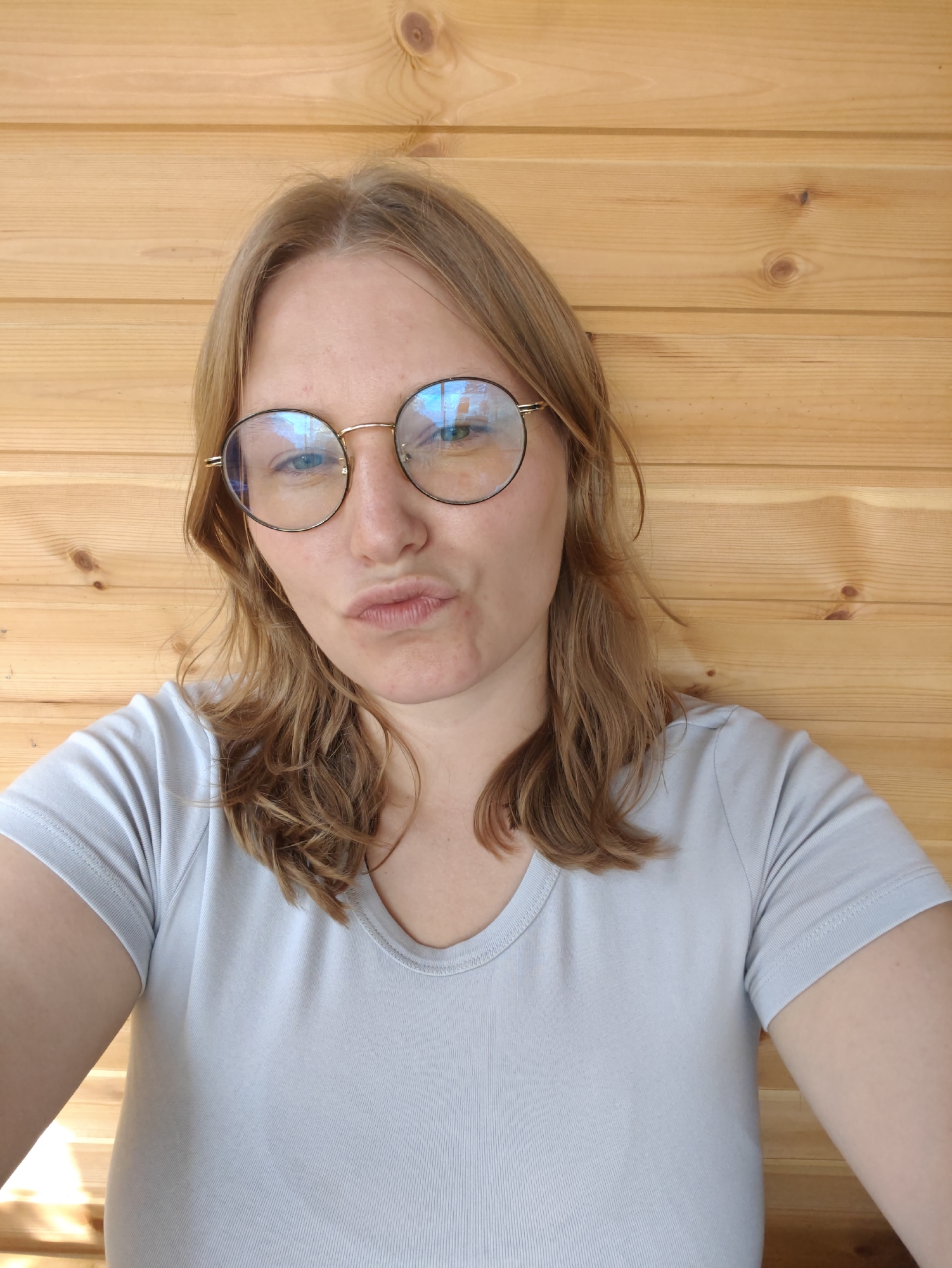 A woman with blonde hair and glasses is posing for a selfie in front of a wooden wall. She is wearing a light blue t-shirt and has her lips pursed. The wooden wall appears to be made of vertical planks. The lighting suggests an indoor setting with natural light coming from the side. The image is a close-up shot, focusing on the woman's face and upper body. There is no indication of the city of Podolsk, Russia in the image.