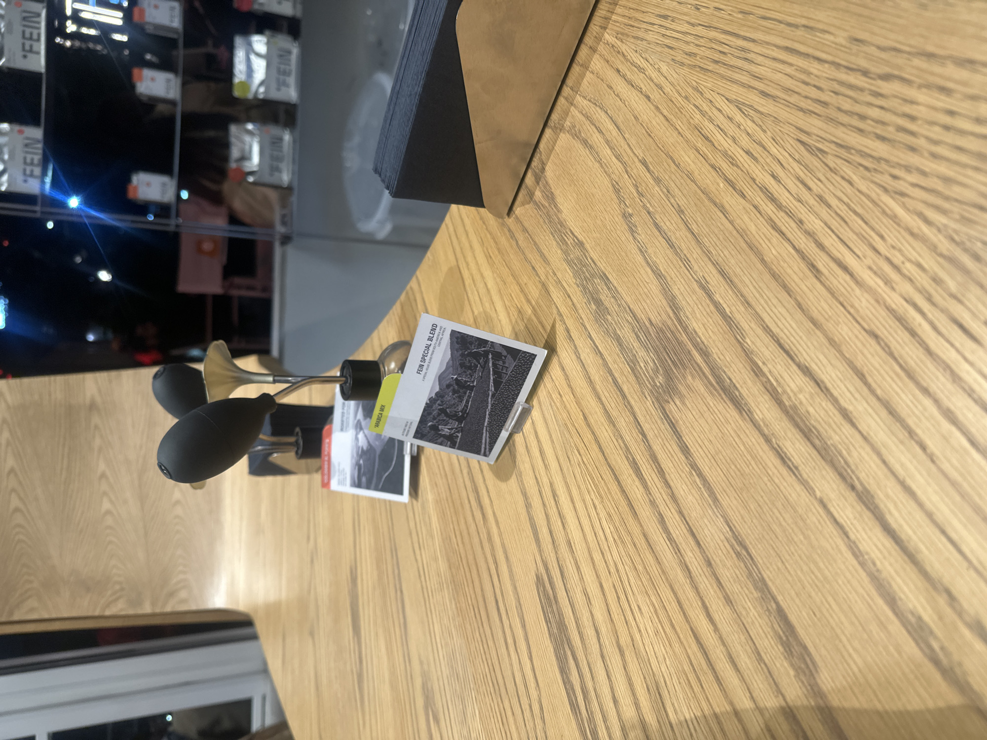 Here is a description of the image:
The scene appears to be an interior shot, possibly within a commercial or exhibition space in Yerevan, Armenia. The foreground is dominated by a light-colored wooden surface with visible grain patterns, likely a table or display counter. Resting on the surface are several items including a black-and-brass device resembling a small, stylized horn or lighting fixture, and two business cards or promotional materials. One card prominently displays the words "FEIN SPECIAL BLEND" and an image of coffee beans, suggesting a connection to a coffee brand or event. The card is adjacent to another that reads "ARABICA MIX" with additional descriptive text. The background is slightly blurred, showing elements of a modern interior with shelves displaying items labeled "FEIN," further suggesting a branding context. The overall setting is clean and well-lit, indicating a professional or curated environment.