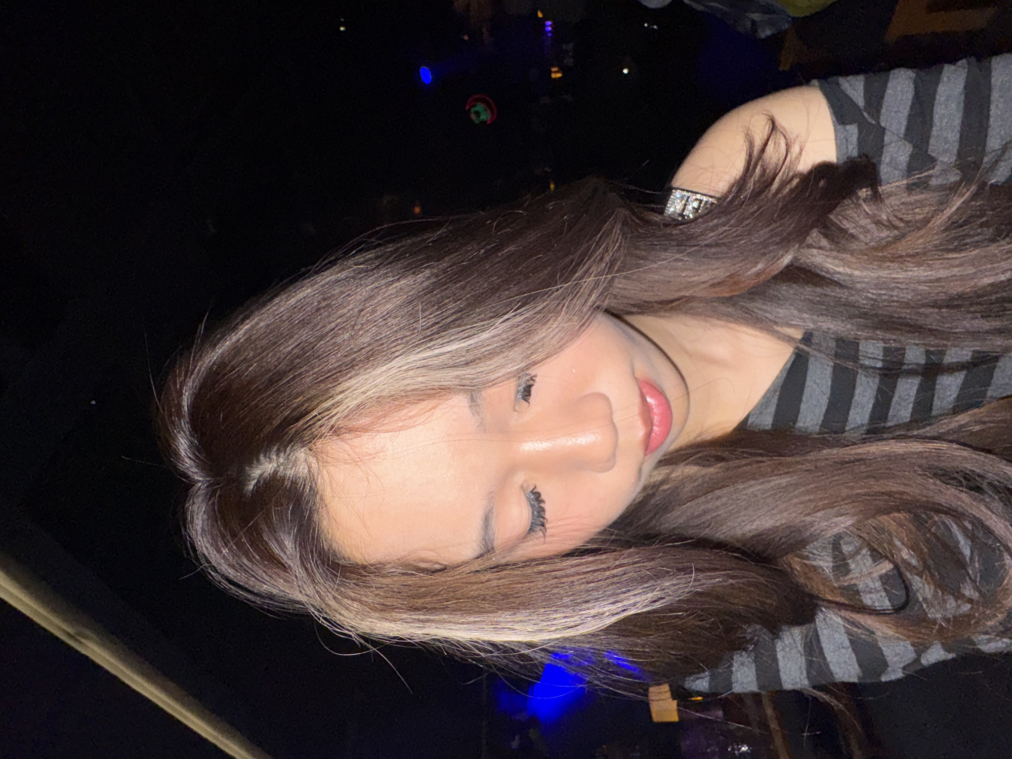 This image depicts a young East Asian female in a dimly lit indoor establishment located in Liangjiang, China. The female, centrally positioned, has long, layered brown hair with prominent lighter highlights framing her face. Her head is tilted slightly forward and downward, with her eyes closed or cast low, and her lips are softly parted. She wears a black and dark gray horizontal striped, off-the-shoulder top. A rectangular, reflective, possibly rhinestone-encrusted embellishment adorns the strap over her left shoulder. Her visible facial features include extended dark eyelashes and subtle glitter on her cheekbones. The background is dark and out of focus, showing indistinct blue ambient lighting on the left, a faint diagonal light-colored beam in the upper left quadrant, and a blurred circular object emitting red and green light on the right, consistent with a club or entertainment venue. No explicit actions or direct interactions are visible beyond the subject's contemplative pose.