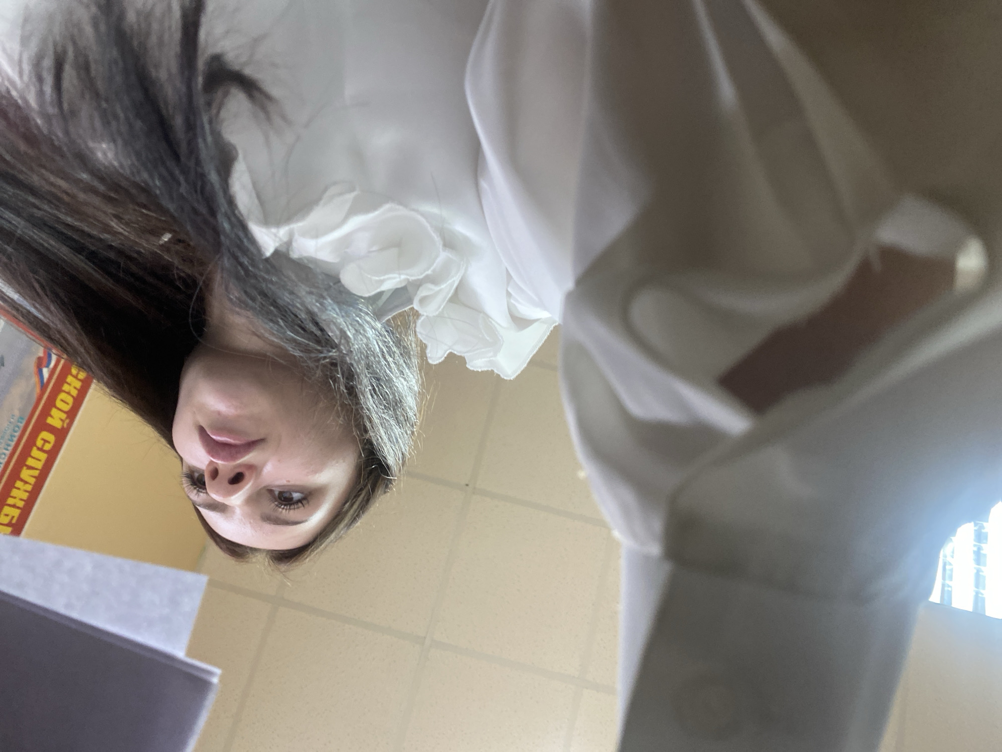 A low-angle, upward-facing photograph captures a young woman from the neck up, wearing a white ruffled blouse. She has long, dark brown hair and is looking down at something out of frame. Her eyes are brown, and she is wearing subtle makeup, including mascara. To her left, a yellow sign with red Cyrillic lettering is partially visible. The background consists of a beige tiled ceiling. A stack of papers or a binder is in the lower left of the frame. The overall impression is that the photo was taken from a very close perspective, looking up at the subject in what appears to be an indoor office or classroom environment. No specific details about the location in Dyakovo, Russia, are discernible from the image.