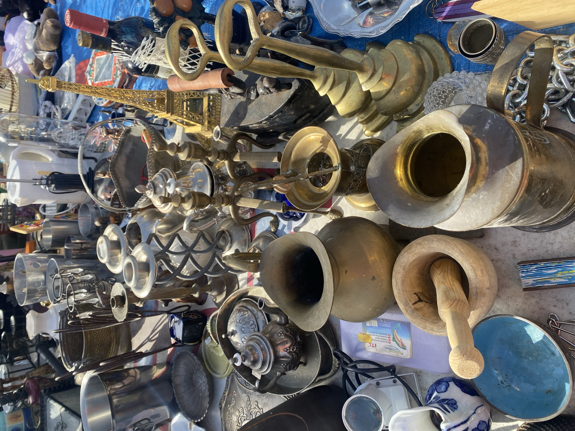 An outdoor market stall in Manilva, Spain, displays a variety of antique and decorative items. The foreground features a large brass pitcher, a wooden mortar and pestle, and a blue ceramic bowl. Behind these, a small silver teapot sits on a tray. The middle ground is occupied by several brass candle holders and a gold Eiffel Tower replica. Further back, various metal containers, bottles, and other assorted objects are visible. The stall is set up on a blue tarp, suggesting a temporary setup for a market. The overall scene conveys a sense of a bustling flea market or antique fair.