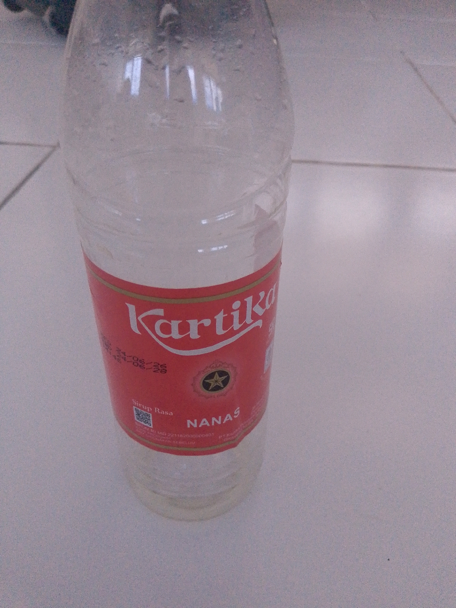 The image shows a close-up of a partially filled plastic bottle of "Kartika" brand syrup. The label is red with white text and features the word "Kartika" prominently. Below "Kartika," it states "Sirup Rasa" and then "Nanas," indicating pineapple flavor. There is a QR code and some text that appears to be regulatory information, including expiration dates. The bottle is sitting on a tiled surface, likely a floor, in what appears to be an indoor environment. The lighting is somewhat dim, and there are reflections on the bottle's surface. The overall scene is static and focuses on the product. There are no people or animals visible, and no indication of the specific location within Putatnganten, Indonesia.