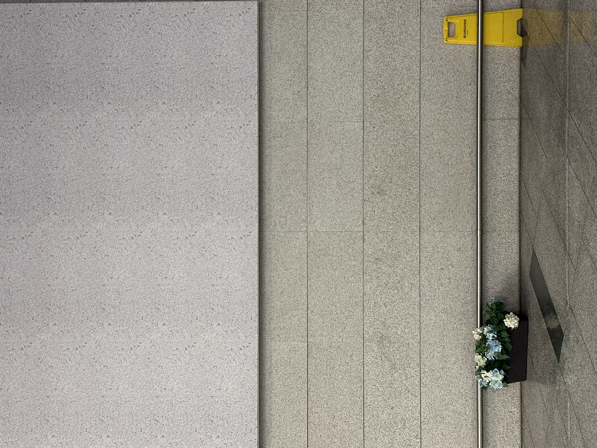 This image presents an indoor setting with tiled flooring and walls, likely a public area. A large gray surface dominates the left side of the image. To the right, the wall is composed of vertical tile panels. A yellow "Caution" sign is placed against the wall, next to a metal handrail. Beneath the rail, there's a planter box with artificial flowers. The floor has a different tile pattern on the right, indicating a transition or change in the space. No people are visible in the scene. The location is unknown.