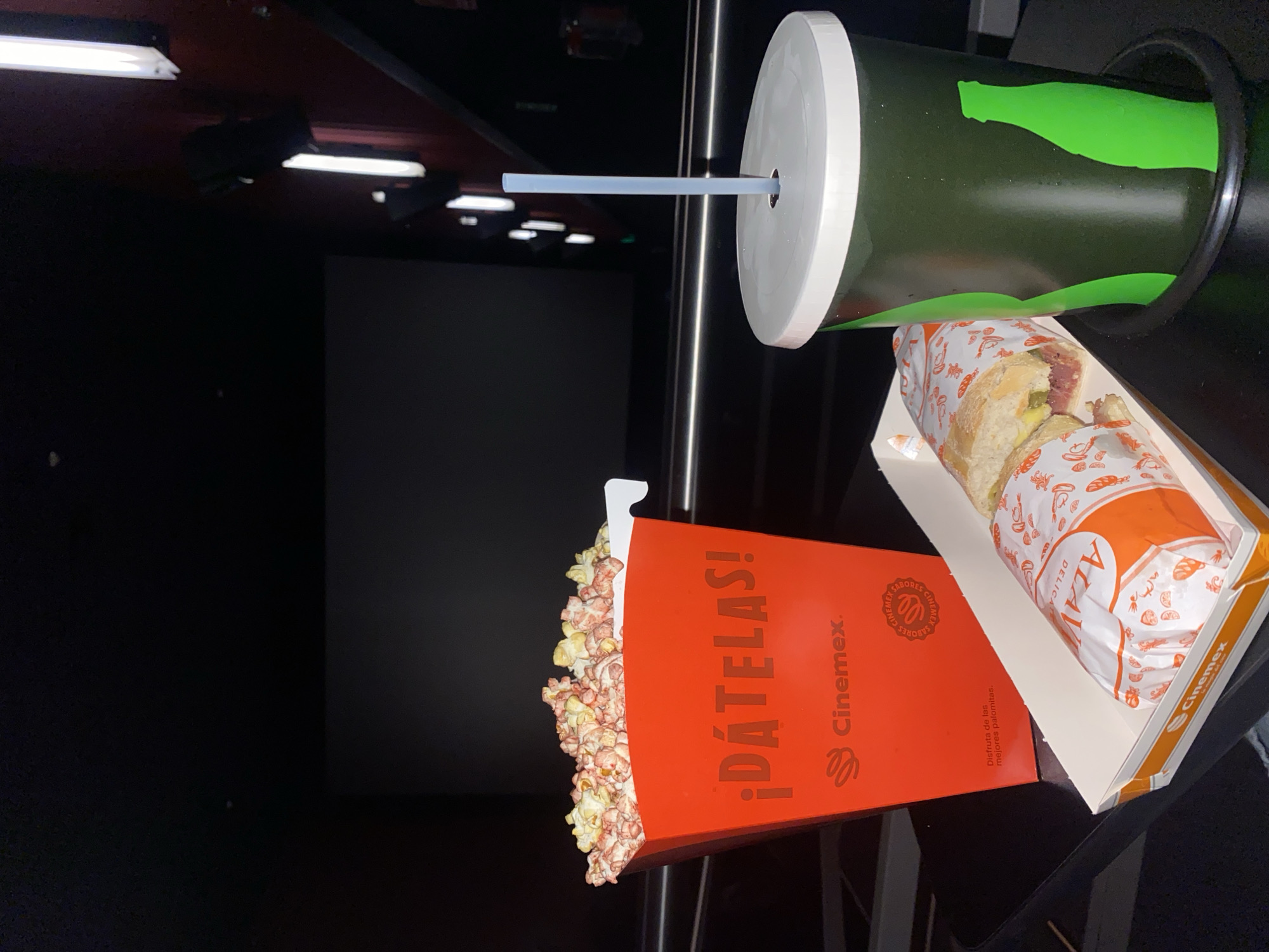 This media file captures a close-up, rotated view of snacks at a movie theater. The image is rotated 90 degrees counter-clockwise, showing items laid out on what appears to be an armrest or small table.
In the foreground, a vibrant red popcorn box is overflowing with popcorn, some kernels spilling onto the surface below. The box prominently features the text "¡DÁTELAS!" and the brand "Cinemex," along with the slogan "Disfruta de las mejores palomitas." (Enjoy the best popcorn). Next to the popcorn, a large, dark green cup with a white lid and a light blue straw is visible. The cup has a bright green, stylized graphic that resembles a bottle or abstract design. Beside the cup is an open, white cardboard tray holding a sandwich wrapped in white and orange paper. The wrapper is patterned with various food items and displays the text "ALAV DELICAT" and "Cinemex." The sandwich appears to be a baguette filled with meat and pickles.
The background of the image is dark, revealing the interior of a cinema hall. A large, rectangular movie screen is visible, currently dark, suggesting the movie has not yet begun or the lights are off. Above, several long, linear fluorescent lights are mounted on a dark-colored ceiling (possibly red or brown), with some of them illuminated. The overall setting indicates an evening or nighttime activity, enjoying concessions at a Cinemex cinema.