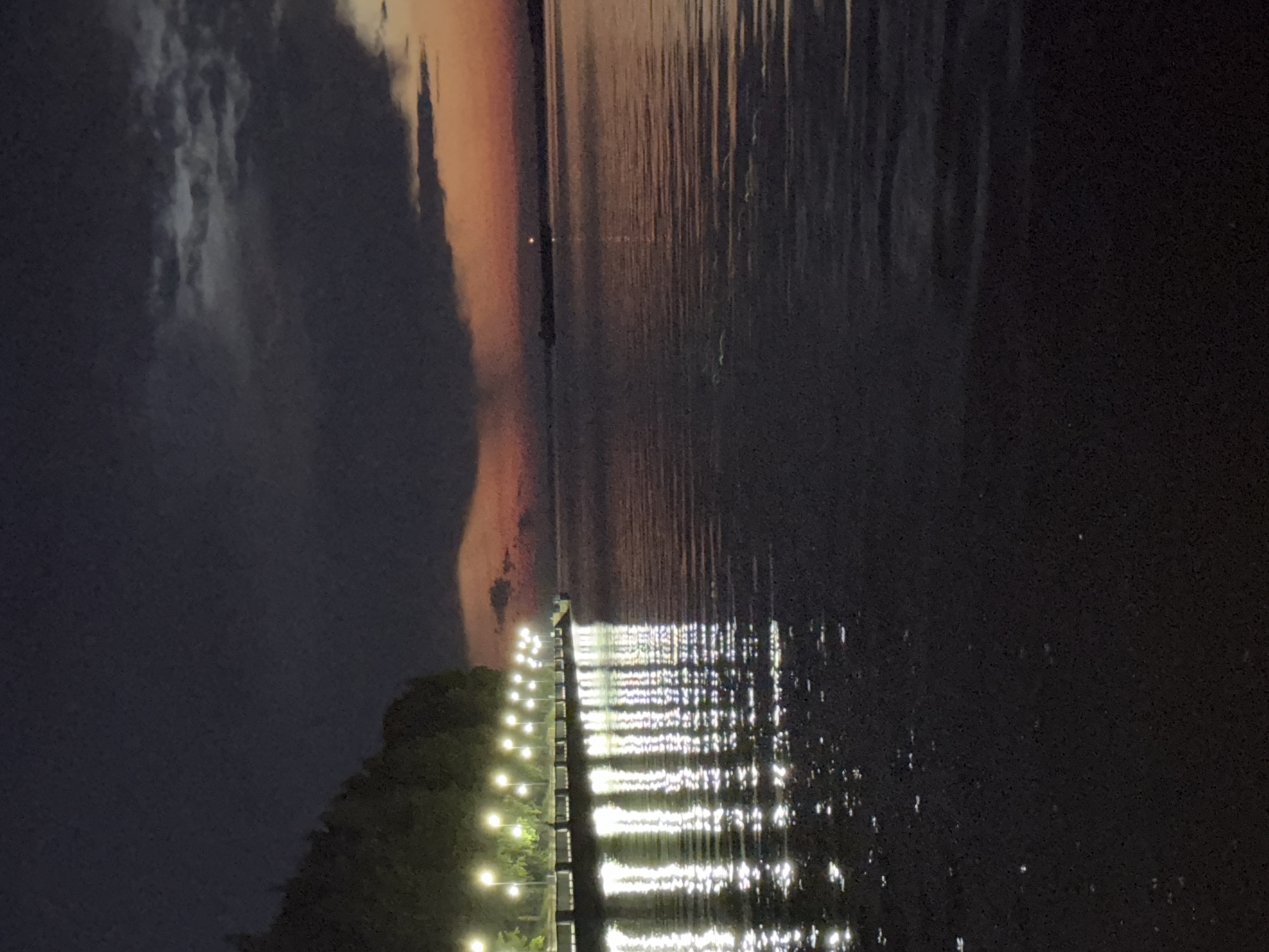 This is a photograph of the embankment in Chaikovsky, Russia, at dusk or twilight. The scene depicts a body of water, likely the Kama River, with a well-lit promenade on the left side. Rows of evenly spaced lampposts illuminate the walkway, casting bright reflections on the water's surface. Trees line the edge of the promenade, their dark silhouettes contrasting with the lights. In the background, the sky displays a gradient of dark clouds at the top, transitioning to hues of orange and pink near the horizon, suggesting the last moments of daylight or the beginning of dawn. The water itself is dark and appears slightly rippled, with the reflections of the lights creating shimmering patterns. The overall atmosphere is calm and serene, highlighting the urban infrastructure against the natural backdrop of the river and sky.