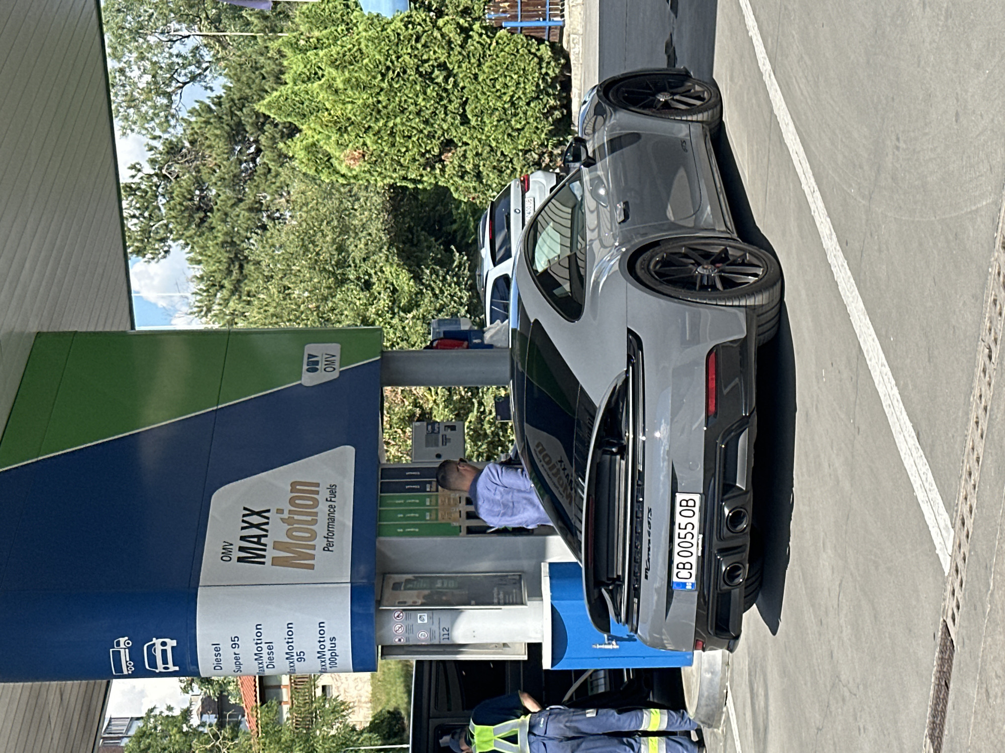A gray Porsche 911 Carrera GTS with Bulgarian license plate CB 0055 OB is parked at an OMV gas station in Veliko Tarnovo, Bulgaria. The car is facing away from the camera, with its rear visible. A fuel pump is next to the car, and a man in a blue shirt and sunglasses is standing by the pump, seemingly refueling. Another person in a yellow vest and blue work pants is standing near a black SUV to the left of the Porsche. The gas station features blue and green signage displaying "OMV MAXX Motion Performance Fuels" and information about different fuel types. The background shows trees and a clear sky.