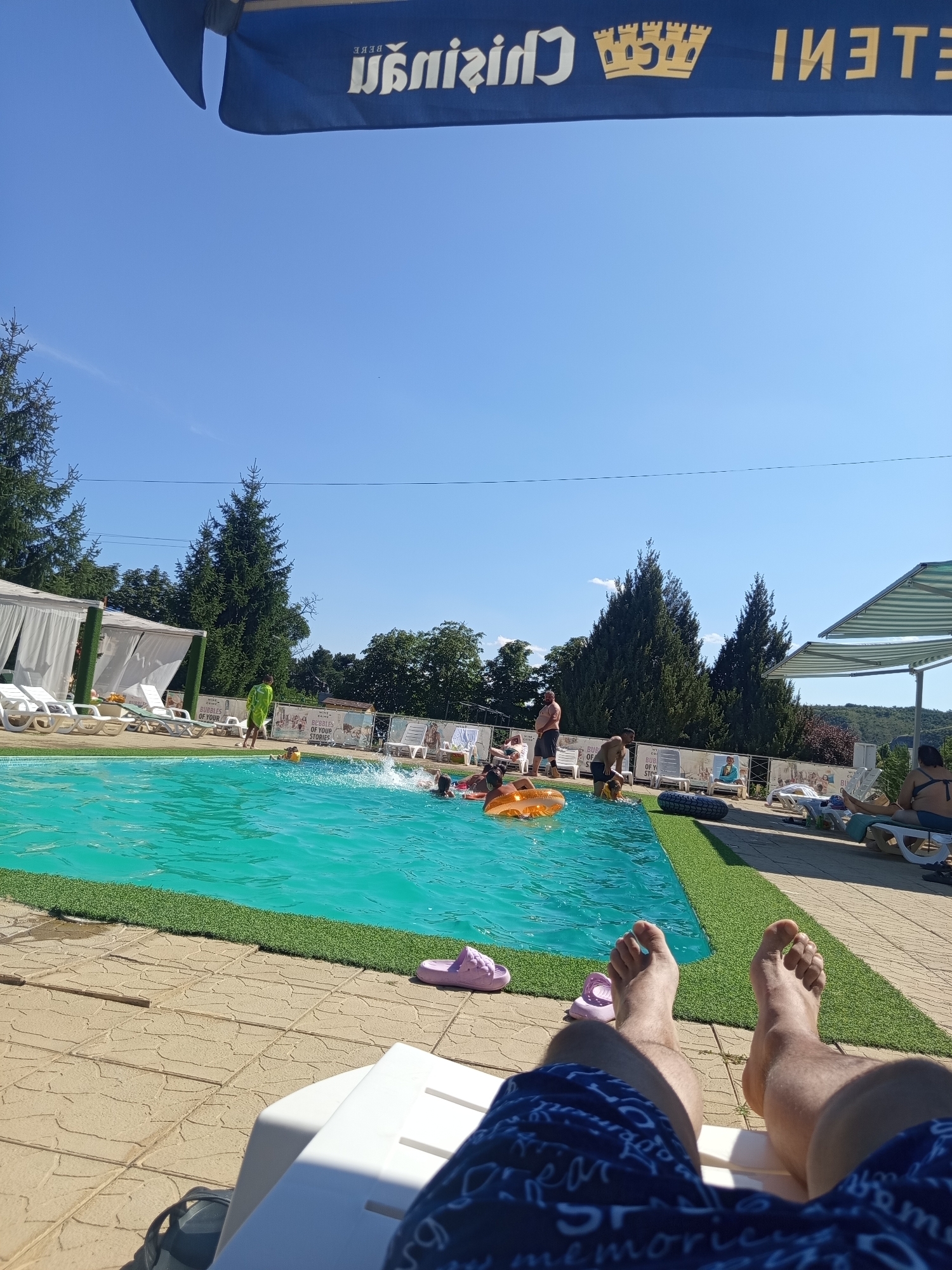 The image depicts a sunny day at an outdoor swimming pool, likely in Molovata Nouă, Moldova, as suggested by external context. In the foreground, a person's legs and feet, clad in blue patterned swim shorts, are visible, resting on a white lounge chair. The feet are bare. To the left and right of the lounge chair, on the light-colored tiled pavement, are two pairs of pink slip-on sandals. A small patch of artificial green turf borders the pool.
The pool itself is filled with bright turquoise water, disturbed by splashes and ripples indicating activity. Several individuals are in the pool, including one person in an orange inflatable ring and others swimming or playing. A person in a bright green garment stands near the edge of the pool, possibly an attendant.
In the background, a white cabana with draped fabric offers shade. Further back, lounge chairs are scattered on the tiled area, some occupied by people relaxing. Trees, predominantly evergreen and deciduous, form a dense backdrop behind the pool area, suggesting a natural or semi-natural environment surrounding the facility. The sky is clear and a bright blue. A blue banner with white lettering, partially obscured by the top edge of the frame, is visible against the sky.